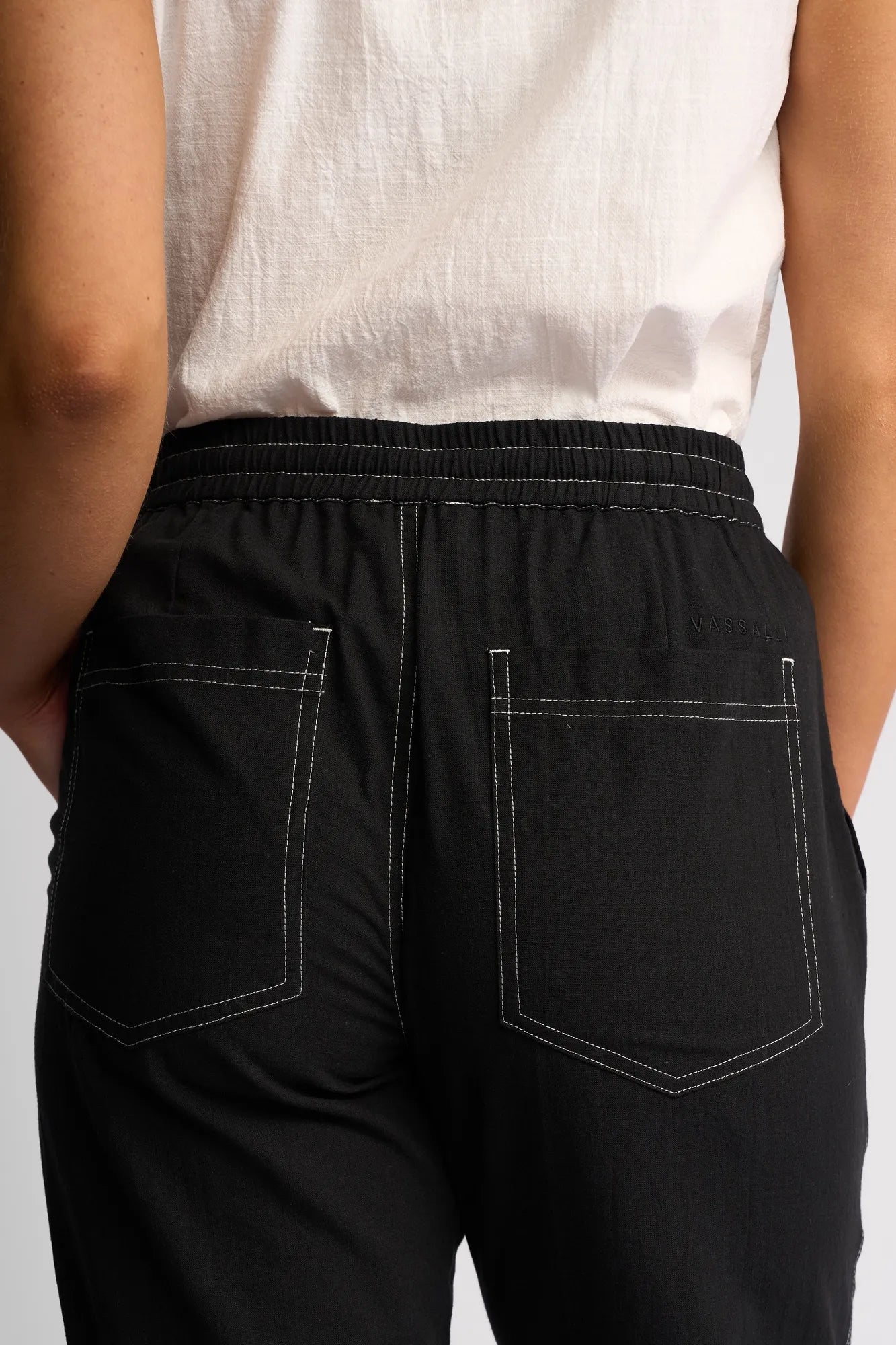 A close-up of a person wearing the Vassalli Tone Pant - Black, styled with white stitching and paired with a white shirt. The image focuses on the back, showcasing two rear pockets and an elastic waistband—a comfortable choice for everyday style.