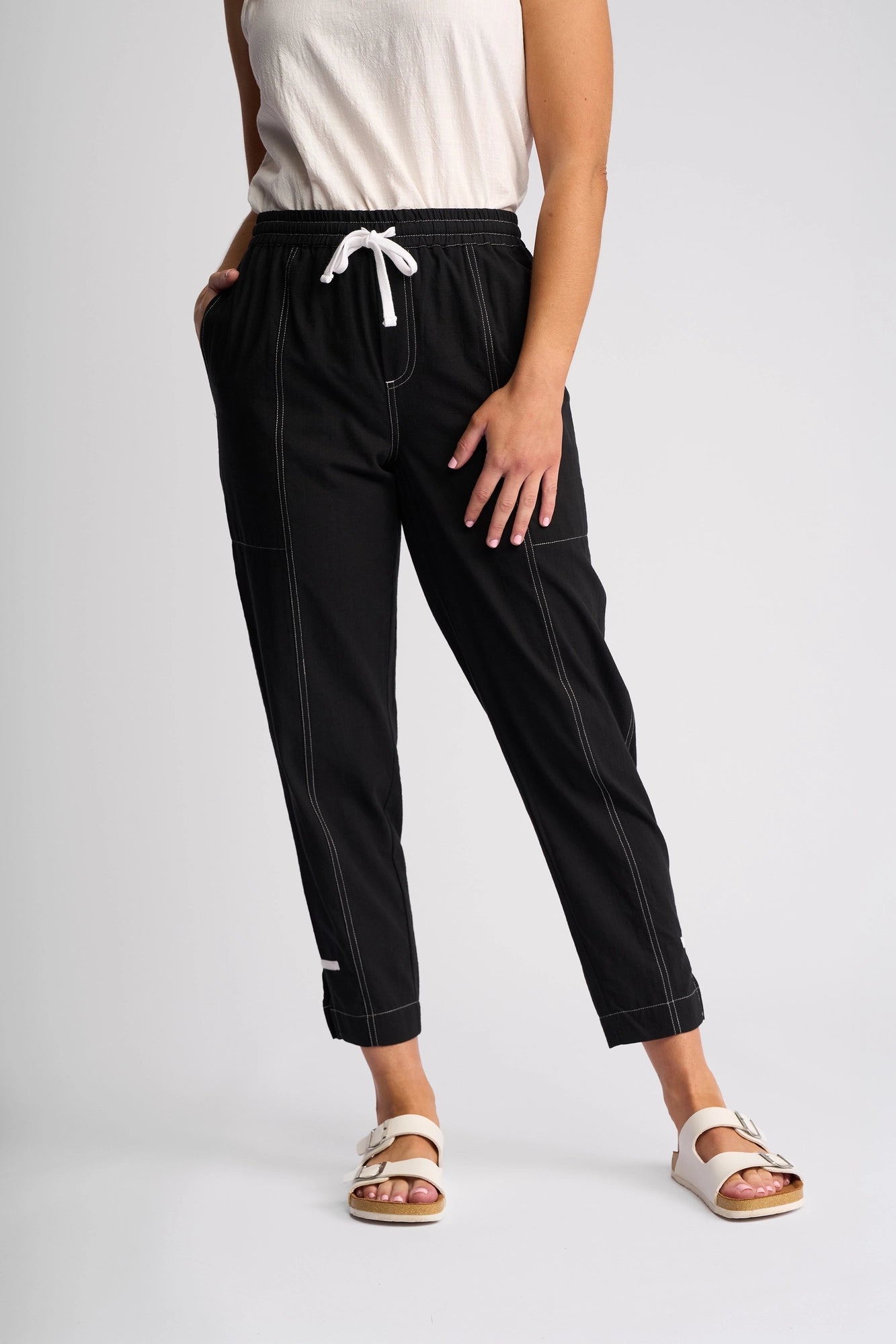 A person stands with hands in pockets against a plain background, wearing the Vassalli Tone Pant - Black featuring white stitching, paired with a sleeveless white top and white sandals—an effortless everyday look.