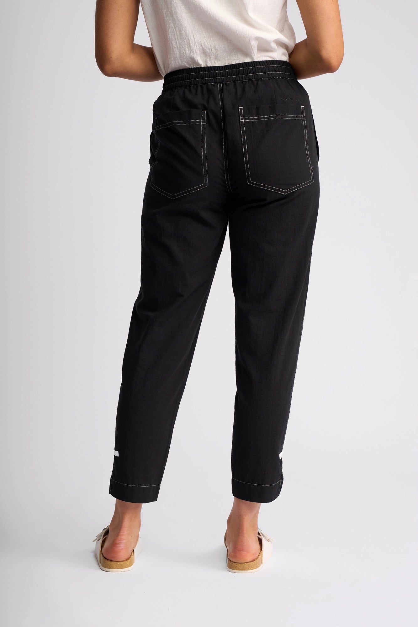 A person stands facing away, dressed in a white shirt paired with the Vassalli Tone Pant – Black, featuring distinctive white stitching for effortless everyday style. Their look is completed with light-colored sandals, and they stand with arms bent in a relaxed pose.