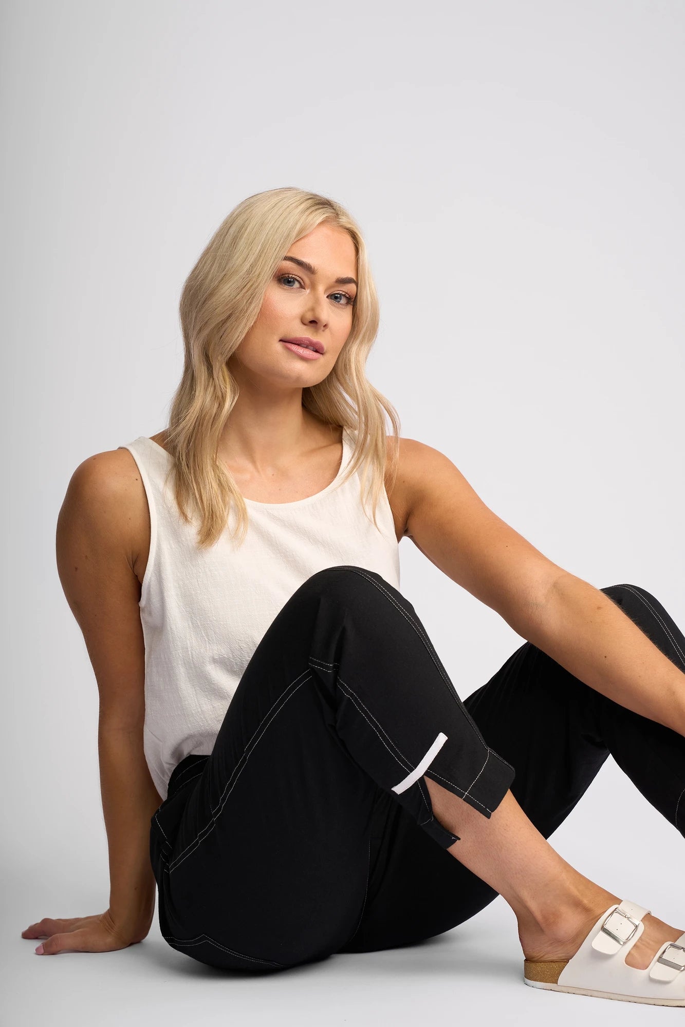 A blonde-haired woman sits on the floor against a light background, wearing a sleeveless white top and the Vassalli Tone Pant in Black, featuring white stitching and crafted from 100% cotton. Finished with beige slip-on sandals, she gazes at the camera with a relaxed expression that highlights her effortless everyday style.