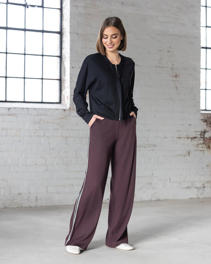 A woman stands indoors by large windows, smiling with her hands in her pockets, wearing the Vassalli Asher Merino Pant in Berry—its relaxed, wide-leg shape makes it perfect for modern everyday wear.