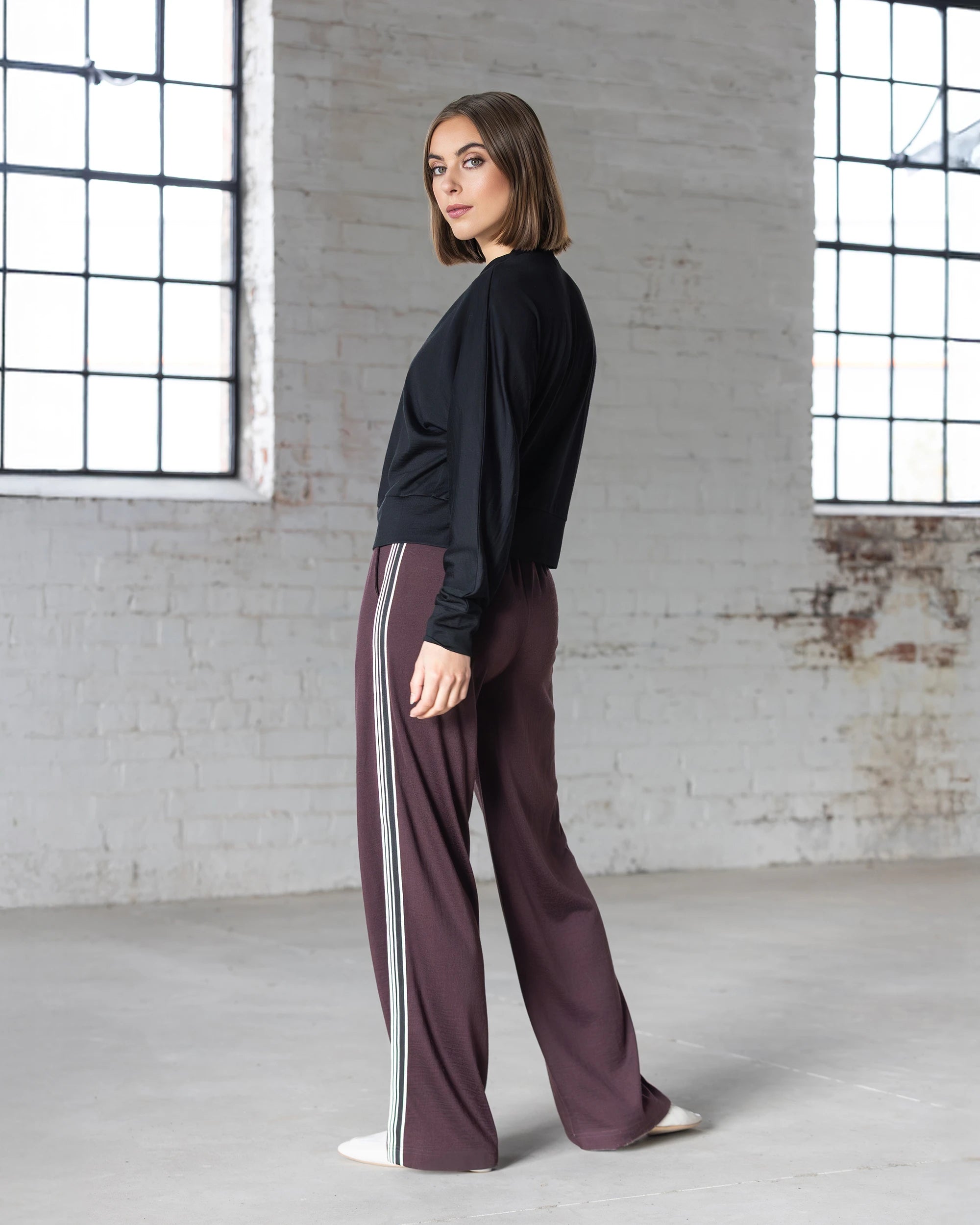 A woman with a bob haircut wears a black long-sleeve top and Vassalli Asher Merino Pant - Berry, featuring white side stripes, as she stands sideways in a bright, industrial-style room—showcasing effortless modern style.