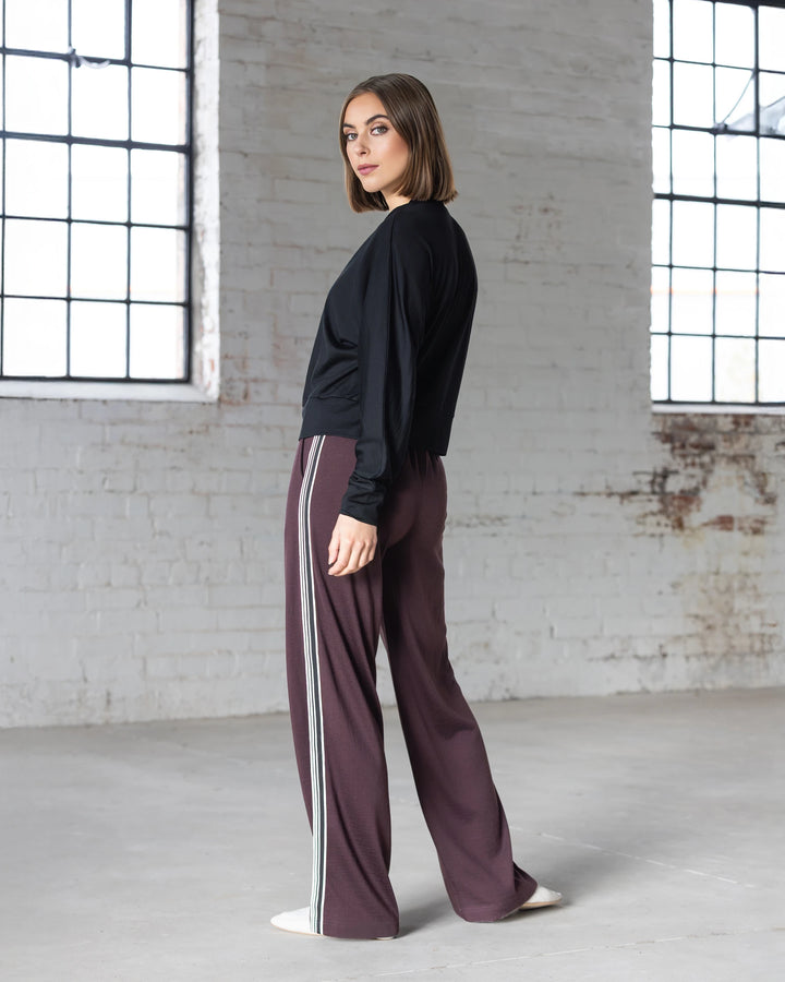 A woman with a bob haircut wears a black long-sleeve top and Vassalli Asher Merino Pant - Berry, featuring white side stripes, as she stands sideways in a bright, industrial-style room—showcasing effortless modern style.