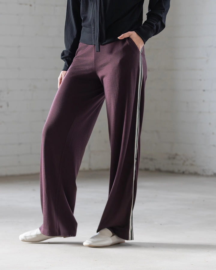 A person stands indoors against a white brick wall, wearing Vassalli's Asher Merino Pant in Berry with a relaxed fit and white side stripe, paired with a black long-sleeve top and white slip-on shoes. Their face is not shown, highlighting modern style.