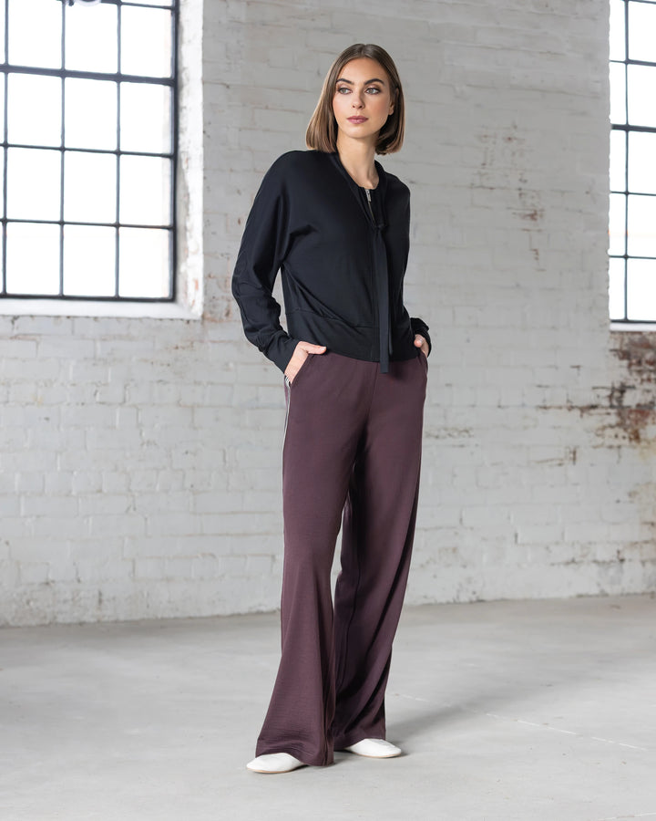 A person stands indoors against a white brick wall, wearing a black long-sleeve top and Vassalli’s Asher Merino Pant in Berry. With hands in pockets and white shoes, two large windows behind them highlight their modern, everyday style.