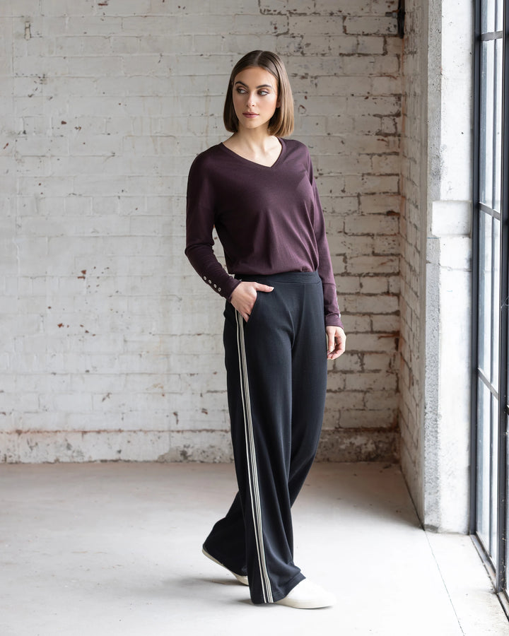 A woman indoors by a large window wears a maroon long-sleeve top and Vassalli Asher Merino Pant - Black, featuring a relaxed wide-leg silhouette with contrast side stripe, paired with white shoes against a white brick wall.