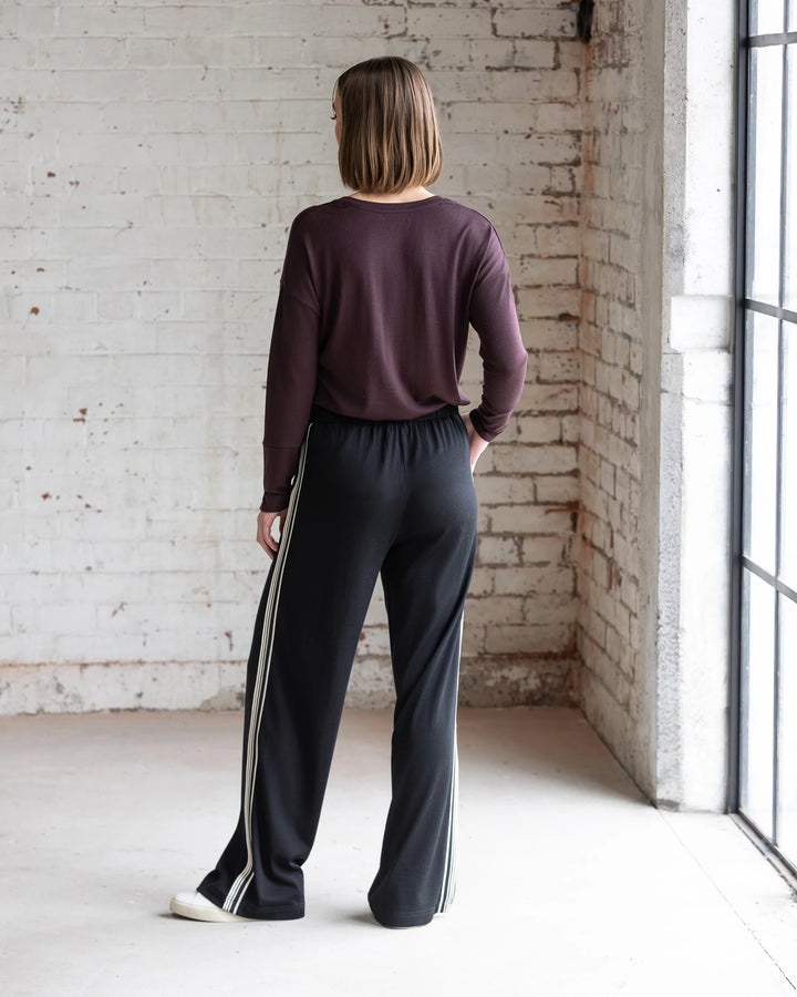 A person with short brown hair stands indoors, wearing a maroon long-sleeve top and Vassalli Asher Merino Pant - Black with a contrast side stripe. Natural light shines in from large windows on the right against a white brick wall.