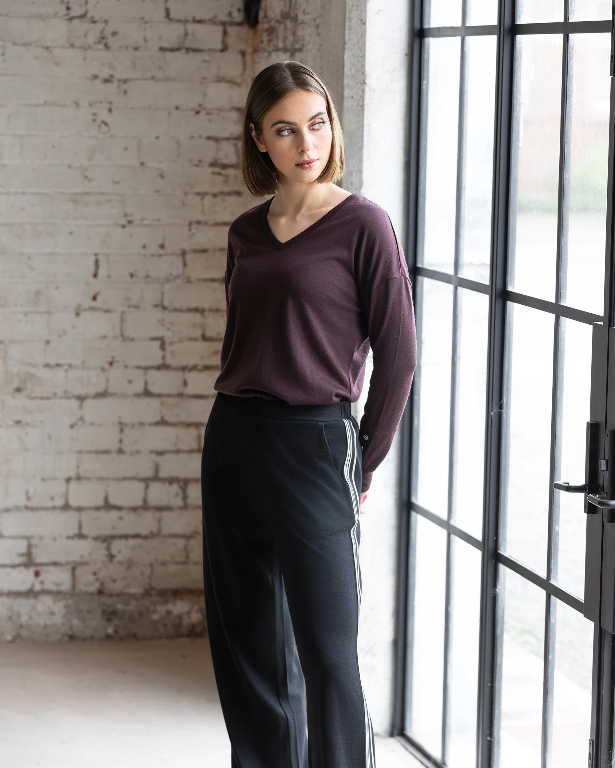 A woman with straight, shoulder-length brown hair stands indoors by a large window, wearing a maroon top and the Vassalli Asher Merino Pant in Black, featuring a relaxed silhouette and contrast side stripe, looking to her left thoughtfully.