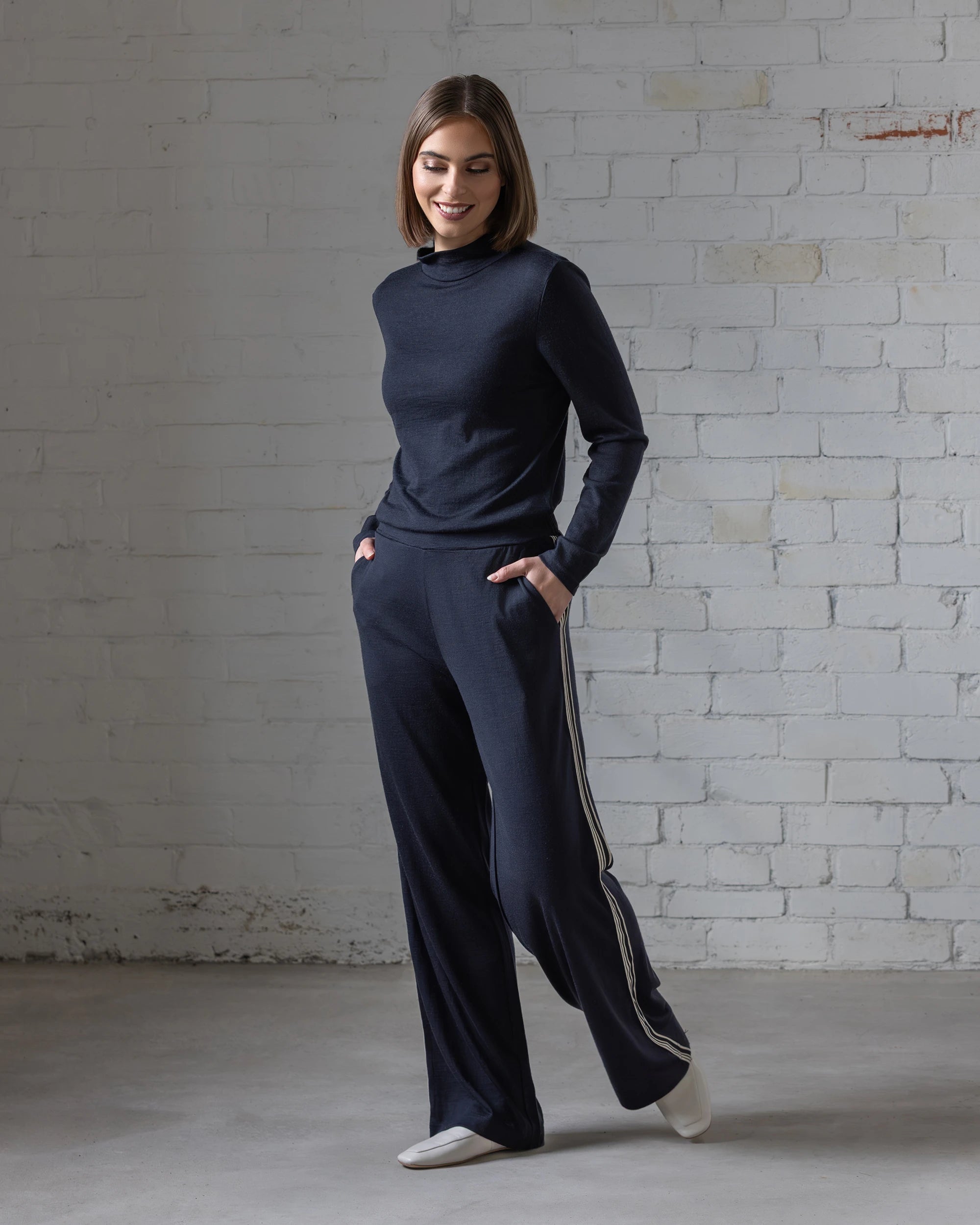 A woman stands indoors by a white brick wall, smiling down with hands in the pockets of her Vassalli Asher Merino Pant - Midnight, featuring white side stripes, paired with a relaxed top and white shoes.