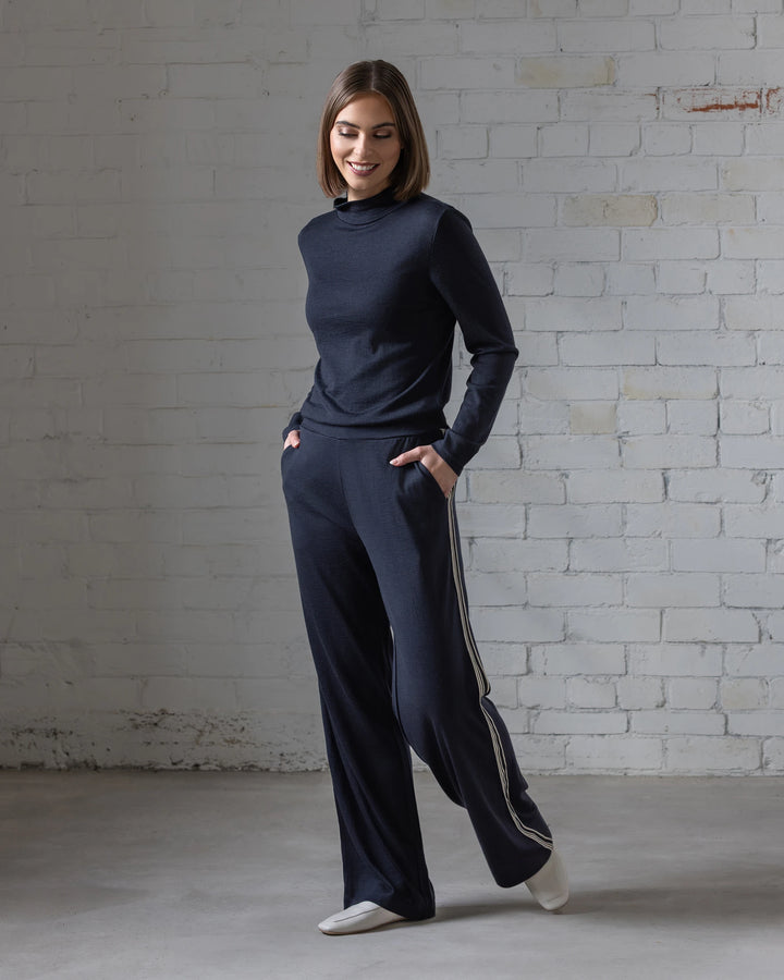 A woman stands indoors by a white brick wall, smiling down with hands in the pockets of her Vassalli Asher Merino Pant - Midnight, featuring white side stripes, paired with a relaxed top and white shoes.
