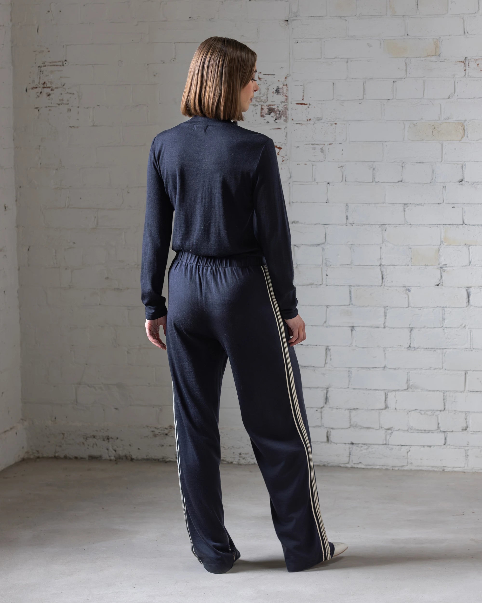A woman with straight, shoulder-length brown hair stands facing a white brick wall, wearing Vassalli's Asher Merino Pant in Midnight—relaxed-fit wide-leg pants with white side stripes—and white shoes. The space features concrete flooring and minimalist decor.