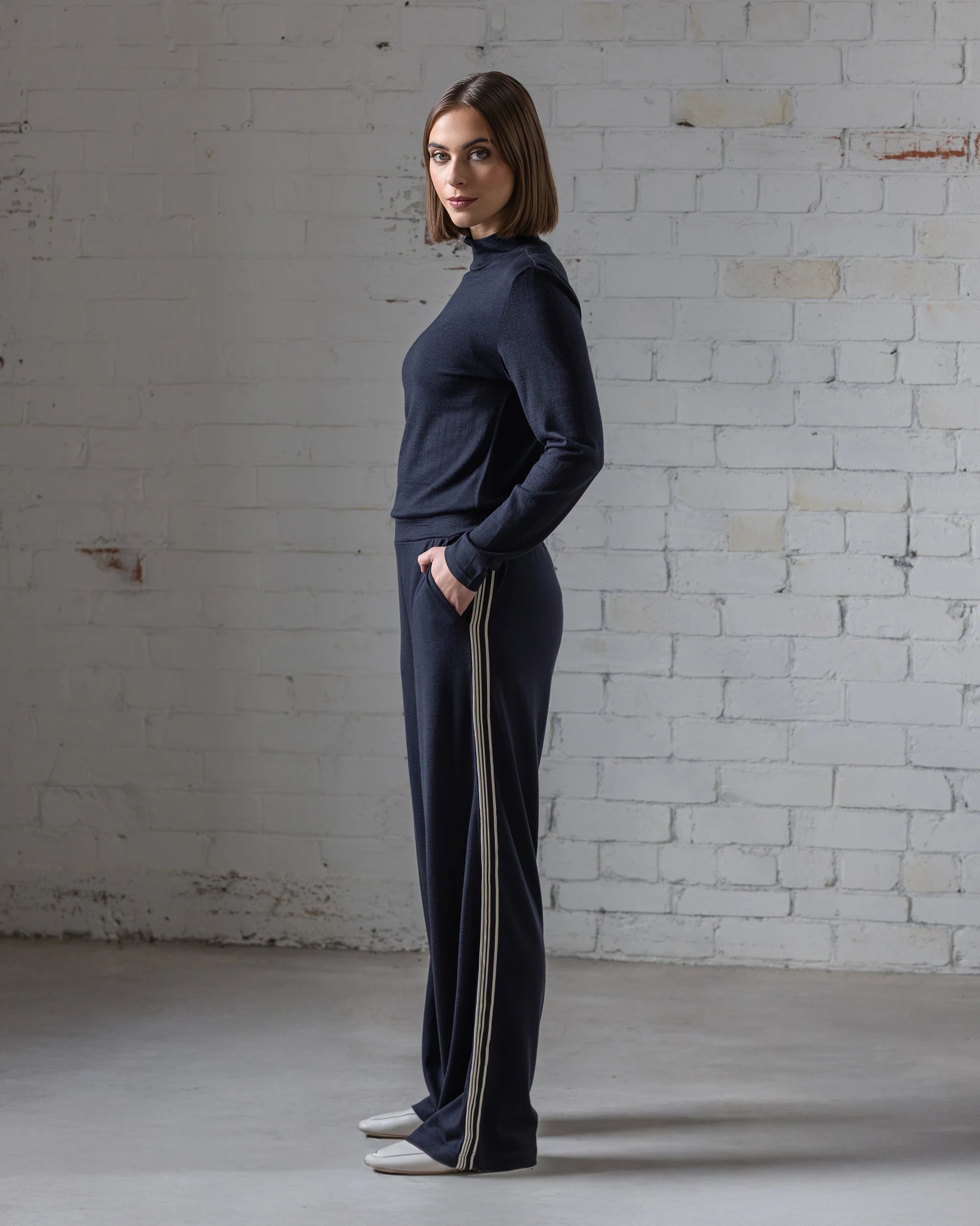 A woman with shoulder-length brown hair stands in profile by a white brick wall, wearing a dark relaxed-fit long-sleeve top and Vassalli Asher Merino Pant - Midnight, featuring wide legs and side stripes. She glances at the camera, looking relaxed.