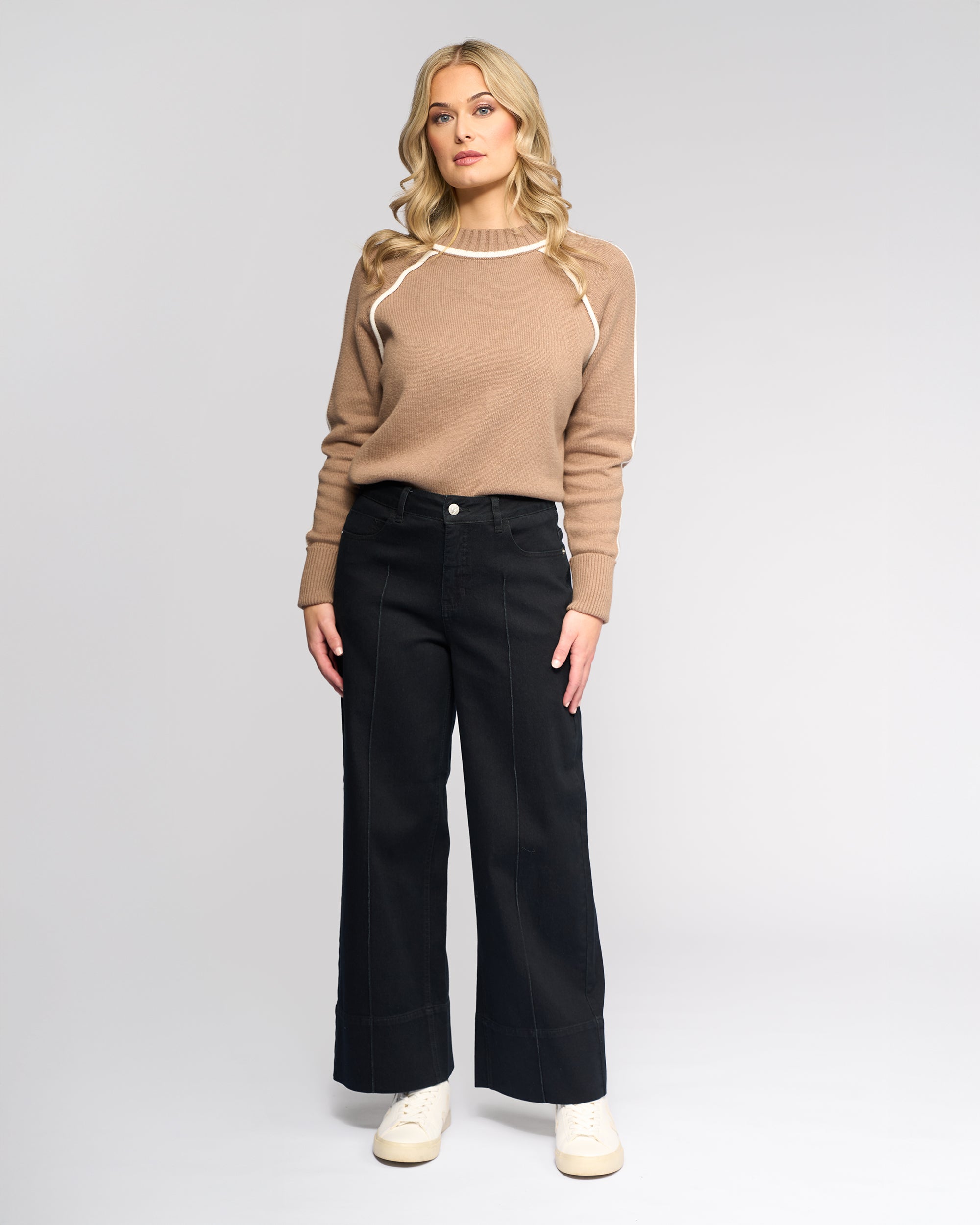 A woman with blonde hair faces forward, wearing Vassalli's Alley Jean in Black Denim, a light brown sweater with white trim, and white sneakers against a plain light gray background.