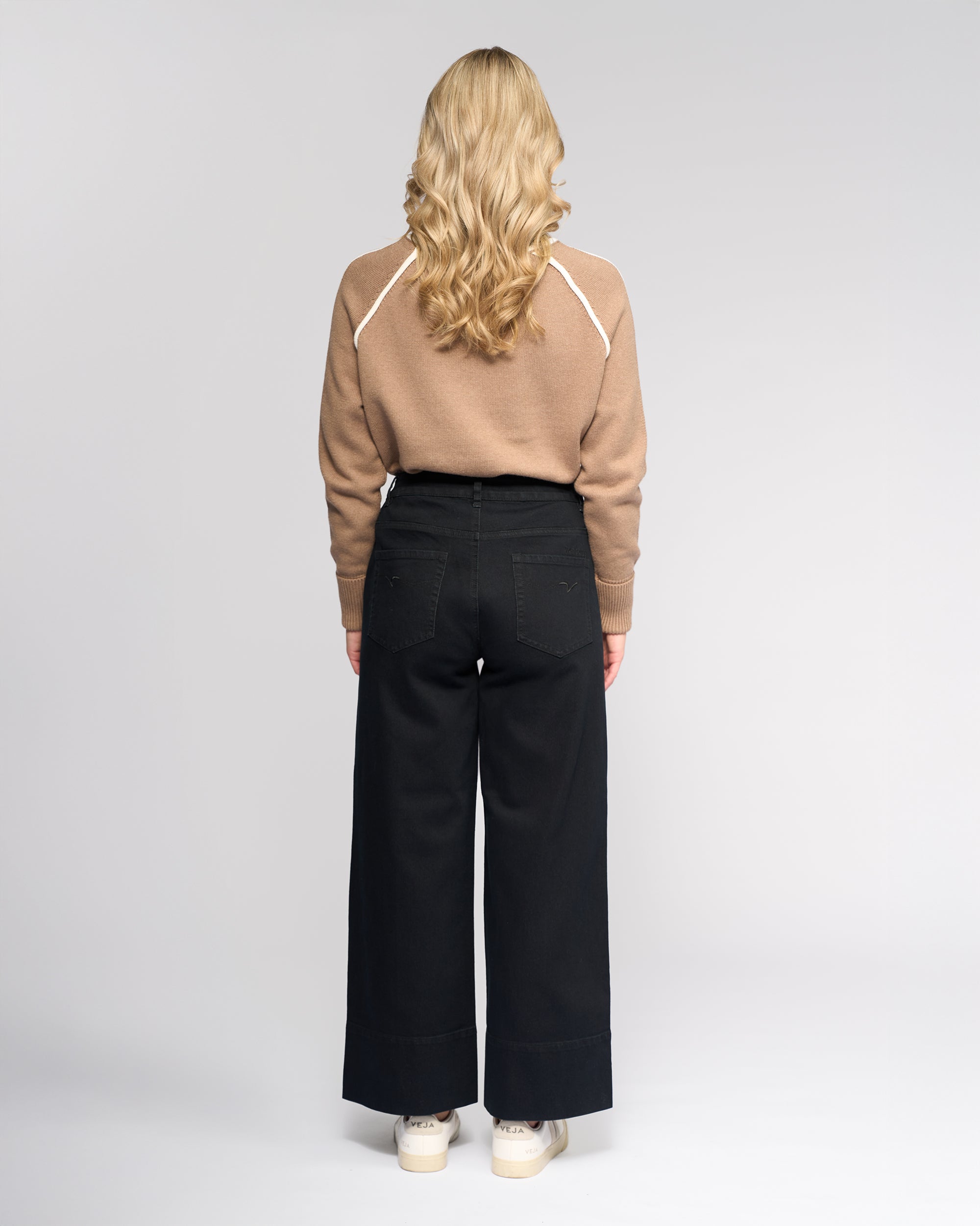 A person with long, wavy blonde hair stands facing away, wearing a tan sweater, white sneakers, and Vassalli Alley Jean - Black Denim against a plain light background.