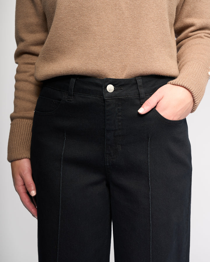A person wearing a beige sweater and Vassalli's Alley Jean - Black Denim stands with one hand in their pocket, showcasing a casual waist-down look.