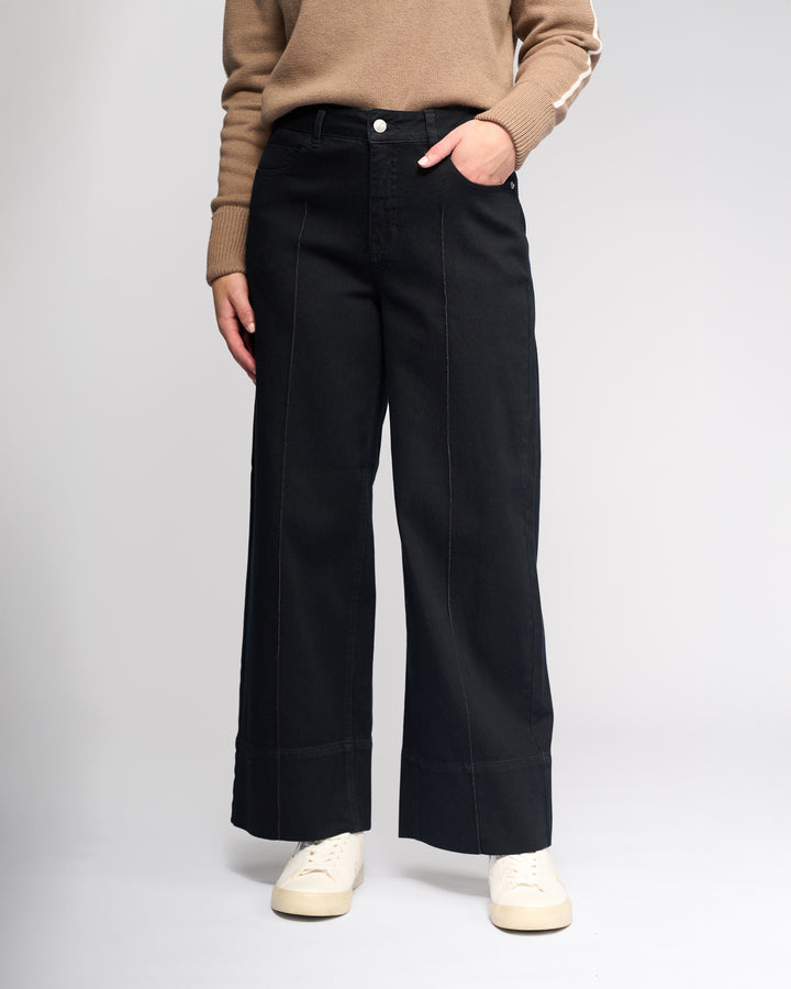 A person stands against a plain light background wearing the Vassalli Alley Jean - Black Denim, a tan sweater, and white sneakers, with one hand in their pocket.