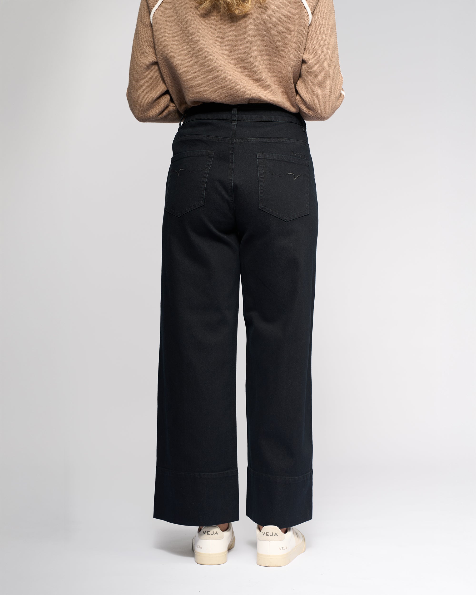 A person stands facing away from the camera, wearing a beige long-sleeve top, Vassalli Alley Jean - Black Denim, and white Veja sneakers against a plain light background.