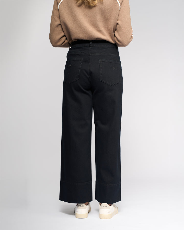 A person stands facing away from the camera, wearing a beige long-sleeve top, Vassalli Alley Jean - Black Denim, and white Veja sneakers against a plain light background.