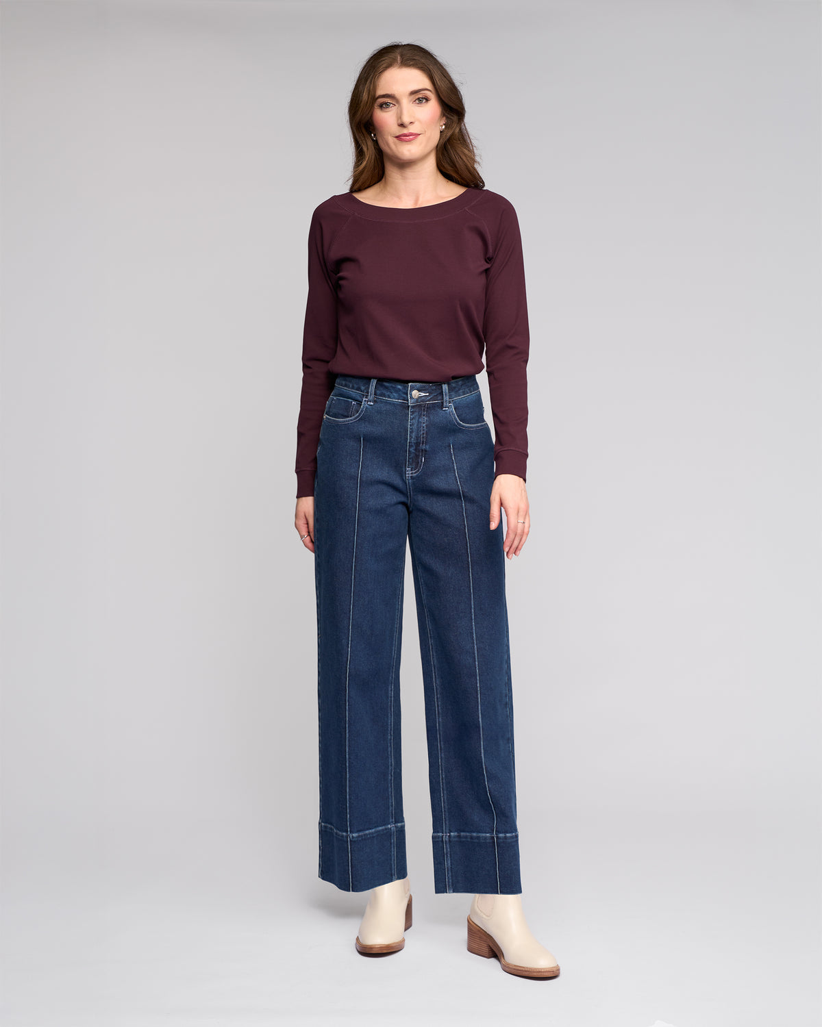 A woman faces forward in a maroon long-sleeve top, Vassalli’s Alley Jean in dark blue with white stitching, and cream ankle boots, set against a plain light gray background.