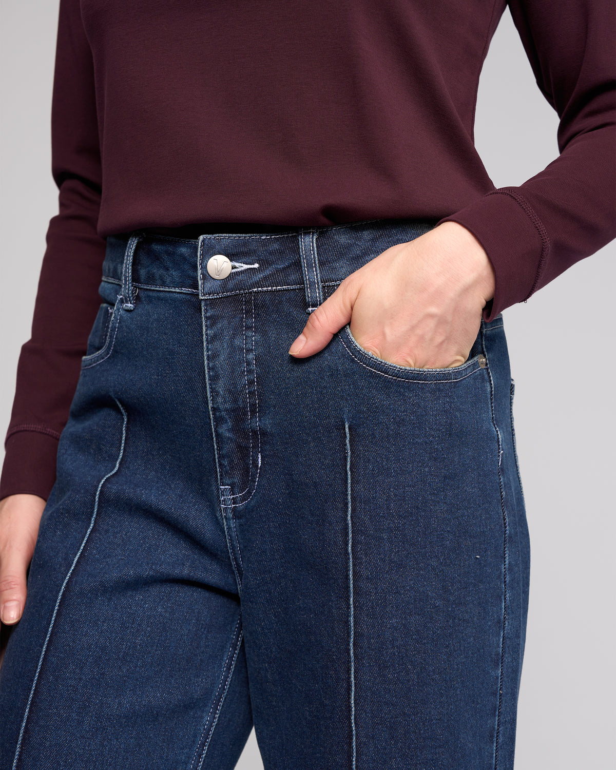A person is shown wearing a burgundy long-sleeve top and Vassalli Alley Jean in dark blue with white stitching, standing with one hand in their pocket. The image displays the torso and upper legs.