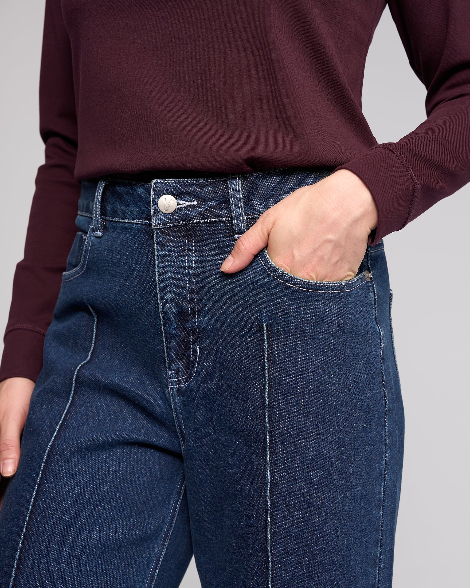 A person is shown wearing a burgundy long-sleeve top and Vassalli Alley Jean in dark blue with white stitching, standing with one hand in their pocket. The image displays the torso and upper legs.