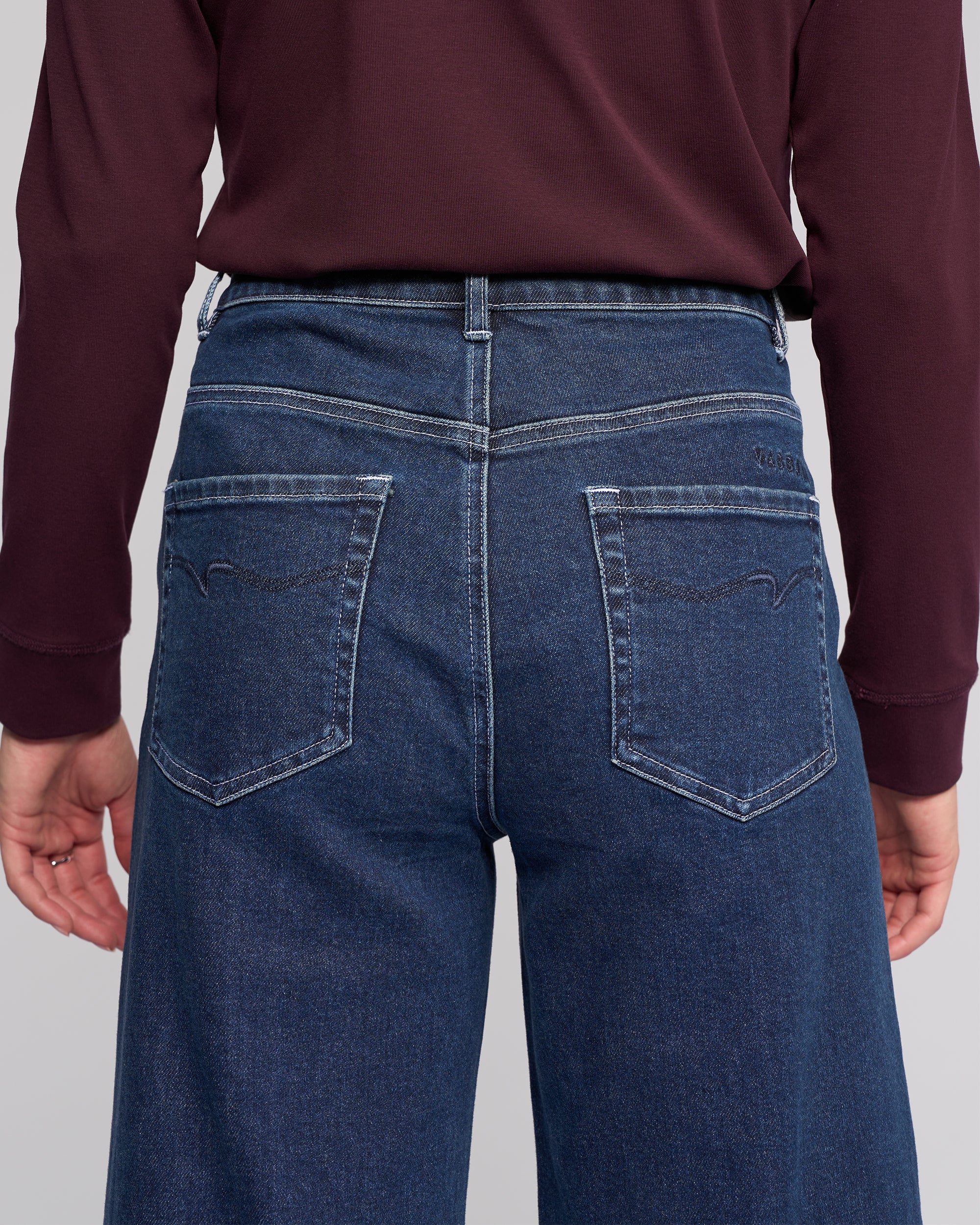 A person is shown from the back wearing a long-sleeve burgundy top and Vassalli Alley Jean - Dark Blue, featuring a high-waisted, wide-leg silhouette, ankle length, and two back pockets with decorative stitching.