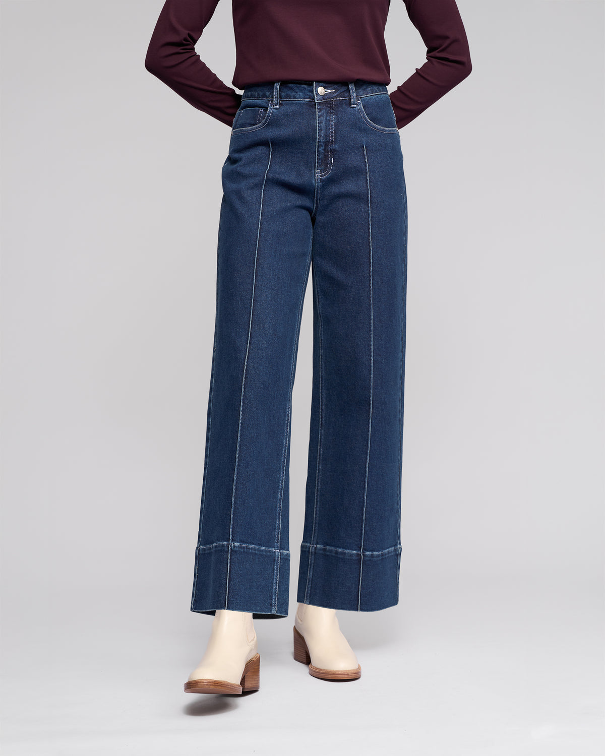 Wearing Vassalli's Alley Jean - Dark Blue, high-waisted wide-leg jeans with front seam details, styled with a long-sleeve burgundy top and white ankle boots. Photo cropped below the shoulders.