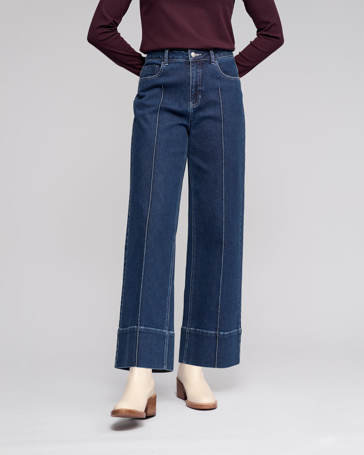 Wearing Vassalli's Alley Jean - Dark Blue, high-waisted wide-leg jeans with front seam details, styled with a long-sleeve burgundy top and white ankle boots. Photo cropped below the shoulders.