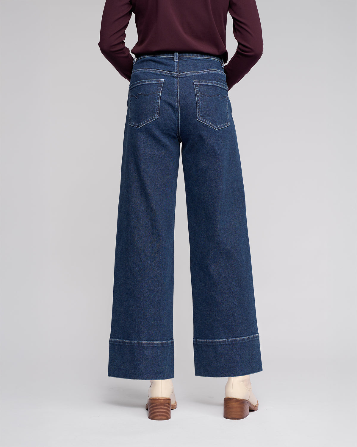 A person faces away from the camera wearing Vassalli's Alley Jean in dark blue, featuring wide-leg, ankle-length style with large back pockets and visible seams, paired with a maroon long-sleeve top and heeled sandals.