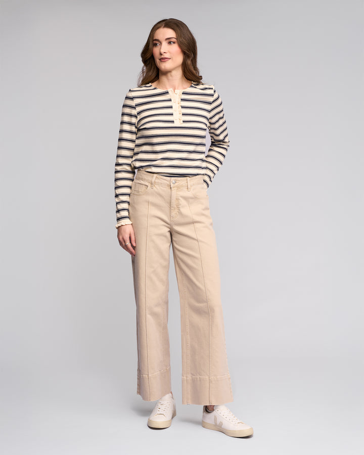 A person stands against a plain background wearing the Vassalli Alley Jean in Oat Denim with a long-sleeve, black-and-white striped top and white sneakers. They have shoulder-length brown hair and strike a relaxed pose with one hand in their pocket.