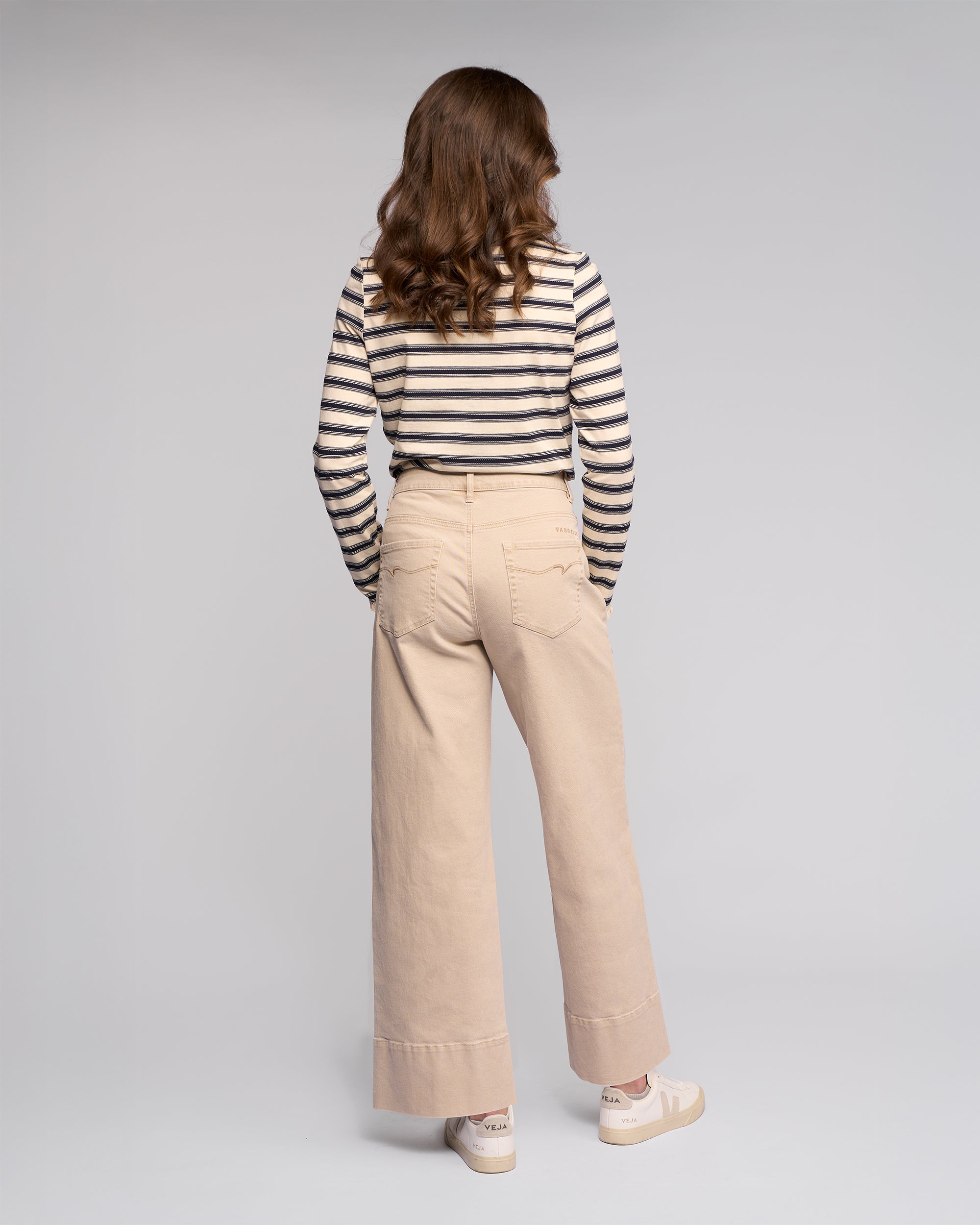 A person with wavy brown hair faces away, wearing a black and white striped long-sleeve shirt, Vassalli's Alley Jean in Oat Denim, and white sneakers against a plain light gray background.