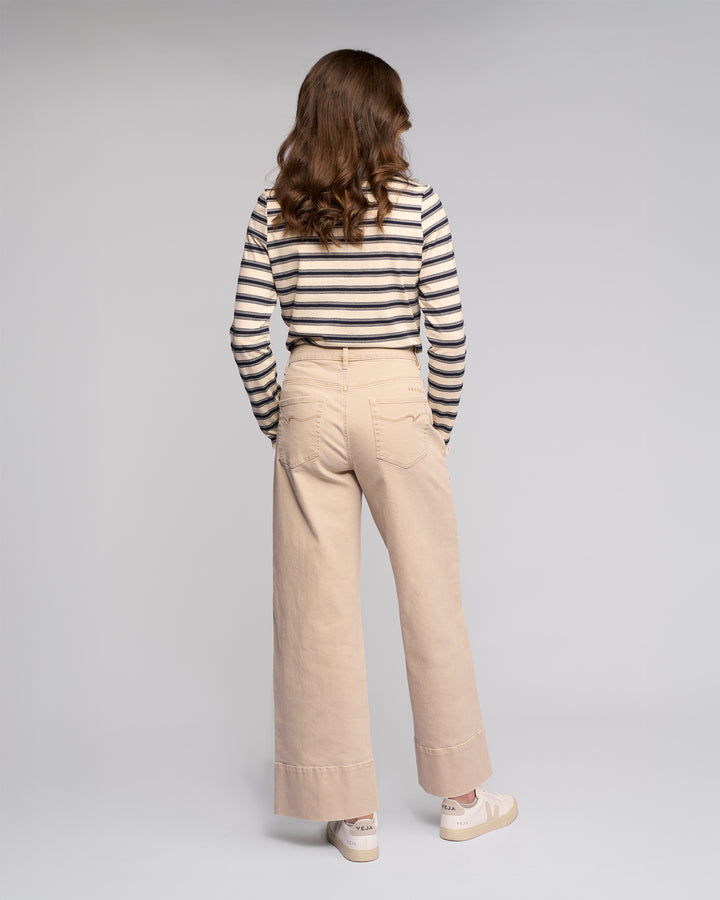A person with wavy brown hair faces away, wearing a black and white striped long-sleeve shirt, Vassalli's Alley Jean in Oat Denim, and white sneakers against a plain light gray background.