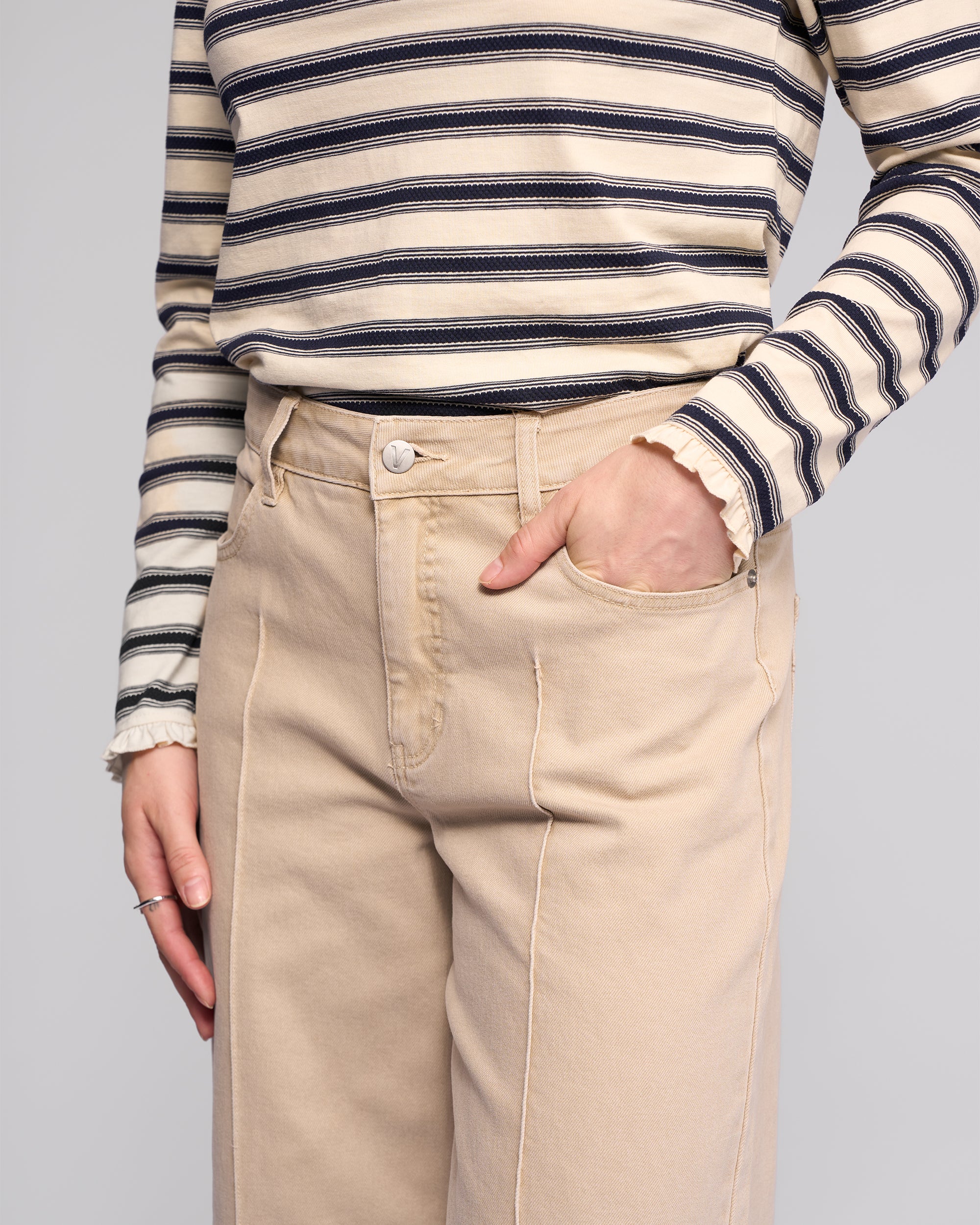 A person wearing Vassalli's Alley Jean in Oat Denim and a beige and black striped long-sleeve shirt stands with one hand in their pocket against a plain background.