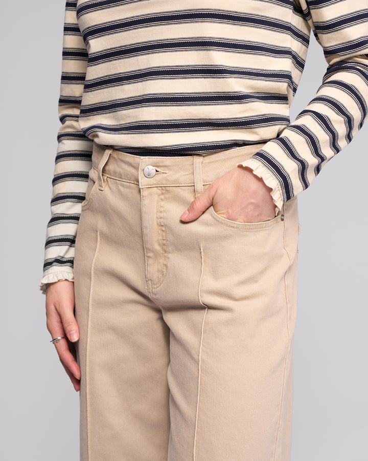 A person wearing Vassalli's Alley Jean in Oat Denim and a beige and black striped long-sleeve shirt stands with one hand in their pocket against a plain background.