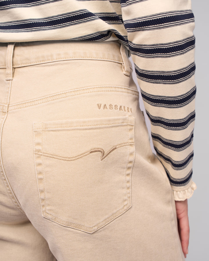 A person wearing Vassalli Alley Jean in Oat Denim, featuring wide legs and signature embroidery above the back pocket, paired with a beige and navy striped long-sleeve shirt; only the waist and upper legs are visible, showcasing a modern silhouette.