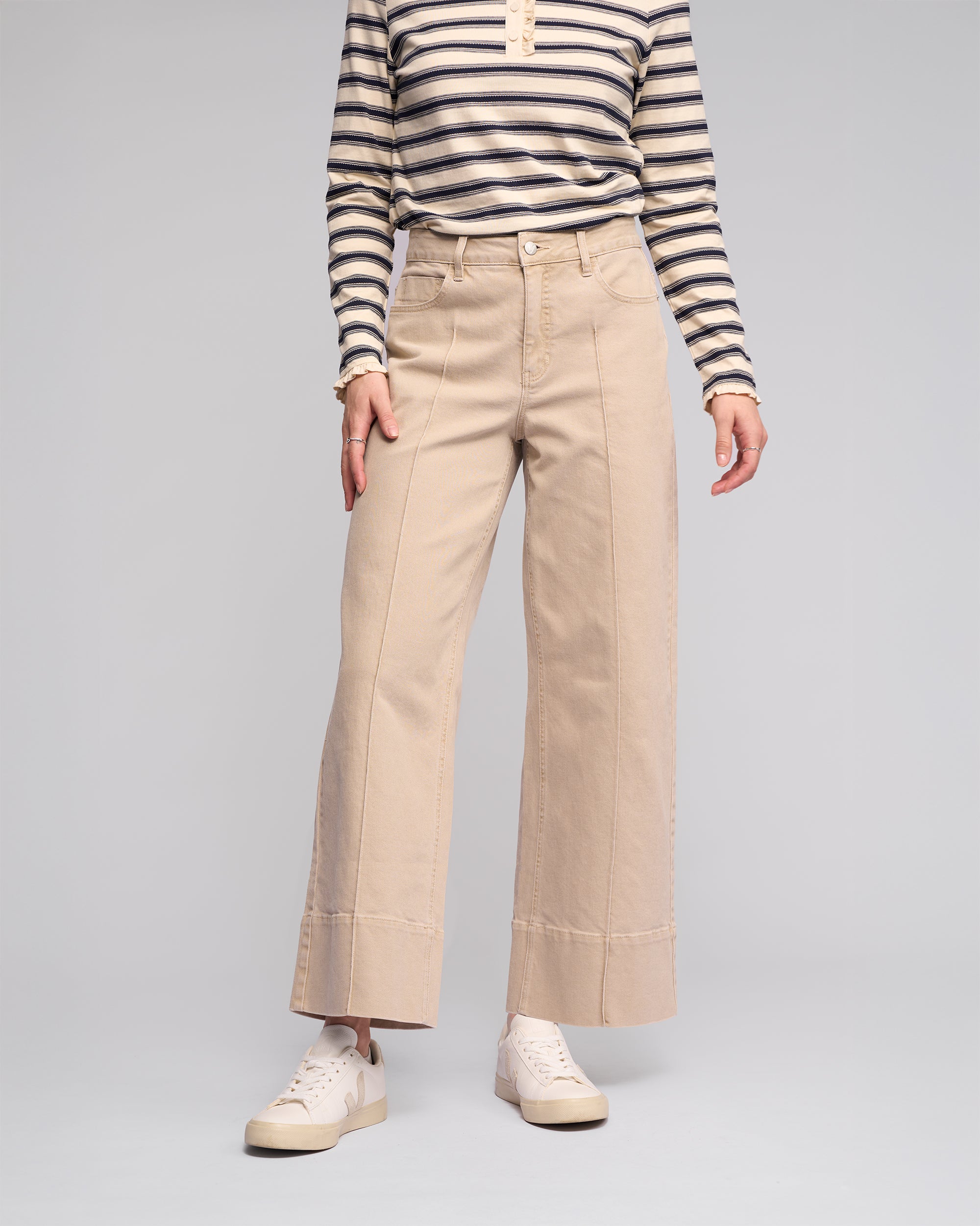 A person models the Vassalli Alley Jean - Oat Denim with visible seams, paired with a long-sleeve black and white striped shirt and white sneakers against a plain gray background. Their face is not shown.