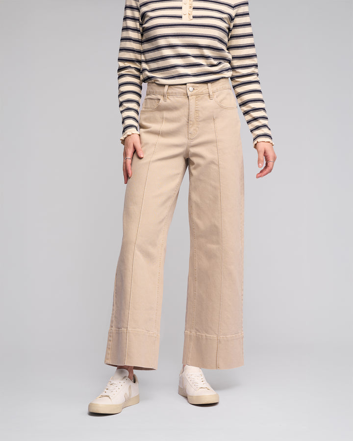 A person models the Vassalli Alley Jean - Oat Denim with visible seams, paired with a long-sleeve black and white striped shirt and white sneakers against a plain gray background. Their face is not shown.