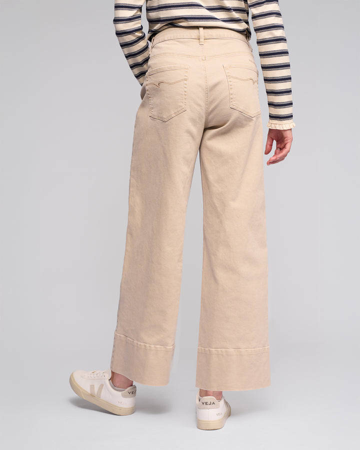 A person shown from the back wears Vassalli's Alley Jean in Oat Denim, styled with a striped long-sleeve shirt and white Veja sneakers, against a plain gray background.