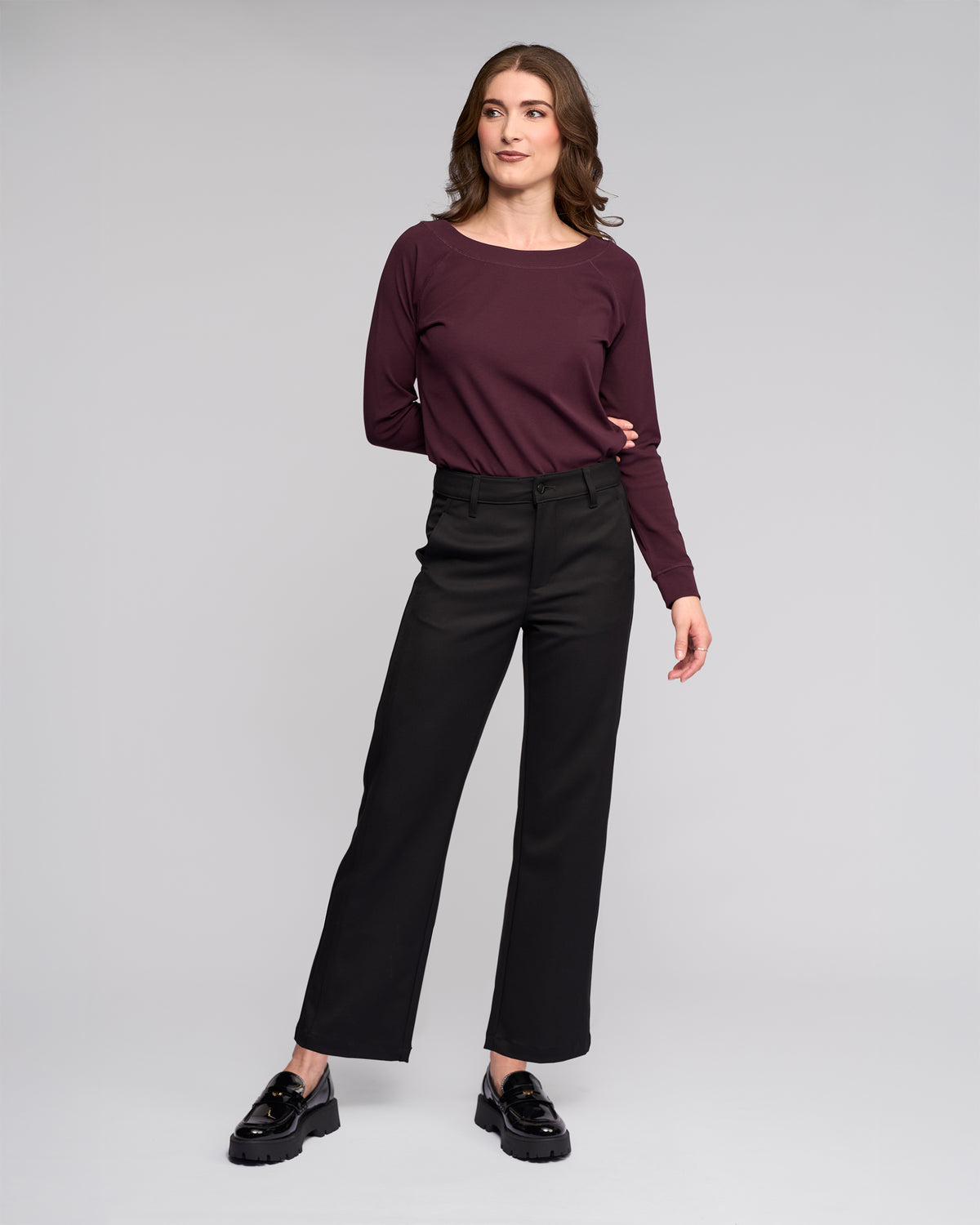 A woman stands against a light gray background, confidently posing in the Vassalli Neve Pant - Black, paired with a maroon long-sleeve top and black loafers.