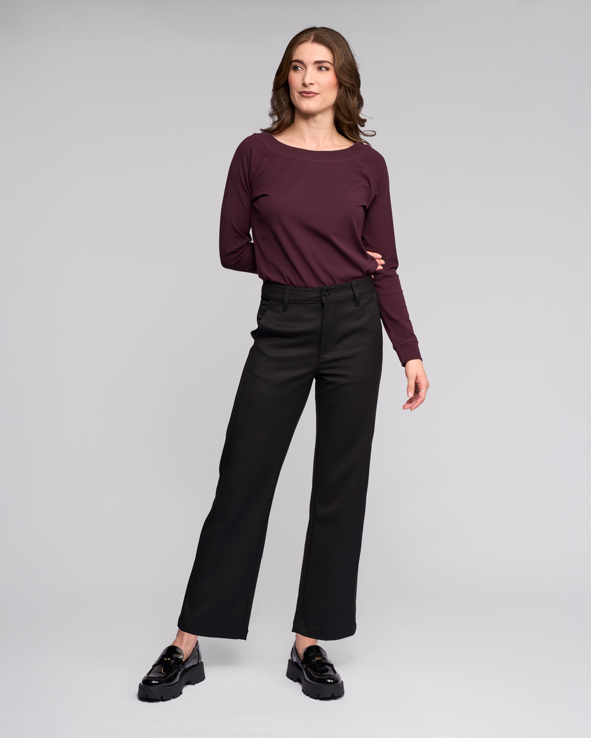 A woman stands against a light gray background, confidently posing in the Vassalli Neve Pant - Black, paired with a maroon long-sleeve top and black loafers.
