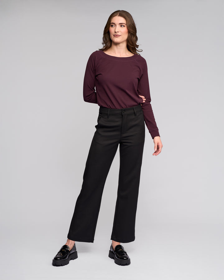 A woman stands against a light gray background, confidently posing in the Vassalli Neve Pant - Black, paired with a maroon long-sleeve top and black loafers.
