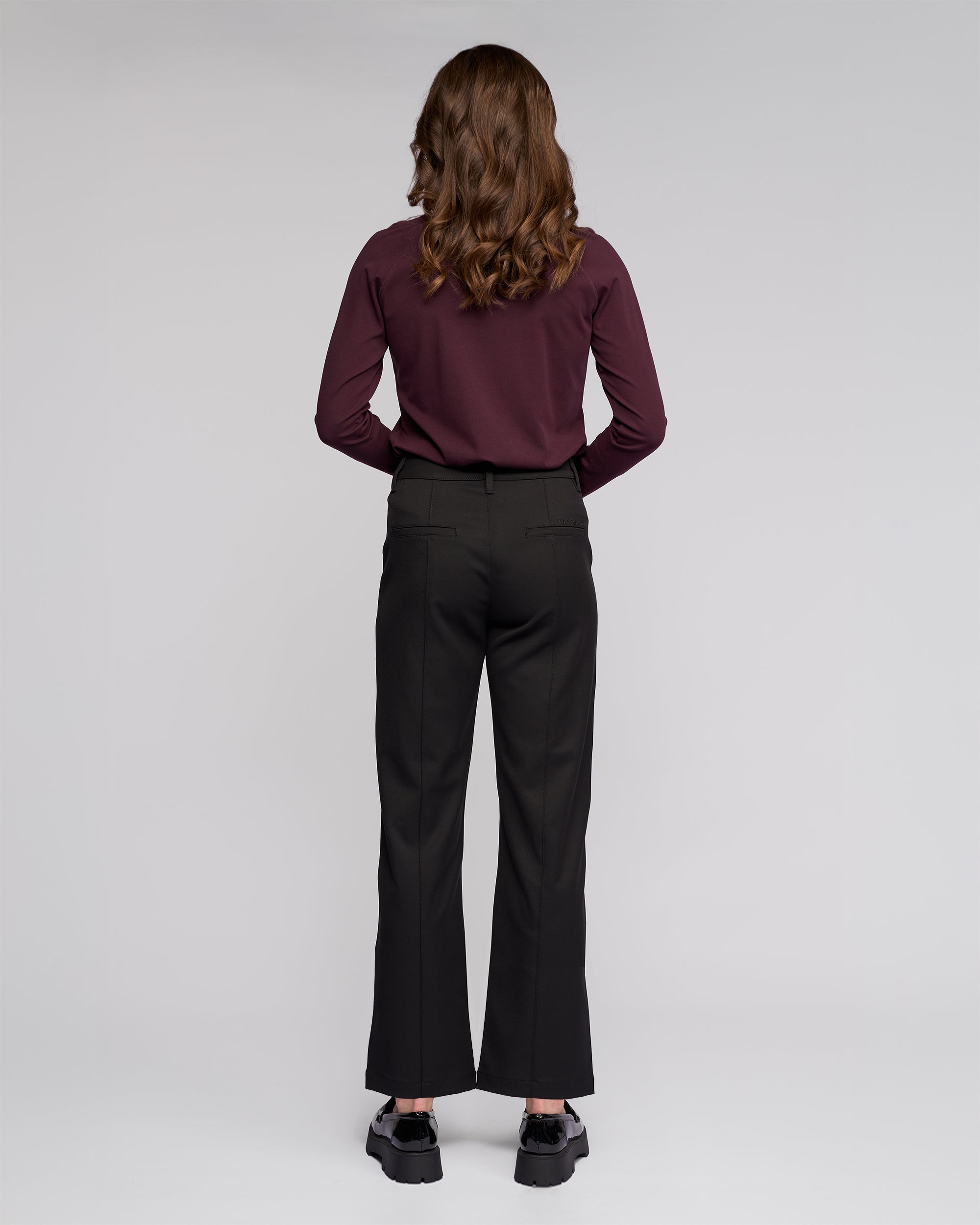 A person with wavy brown hair, dressed in a maroon long-sleeve shirt and Vassalli's Neve Pant in black, paired with black shoes, stands facing away from the camera against a plain light background.