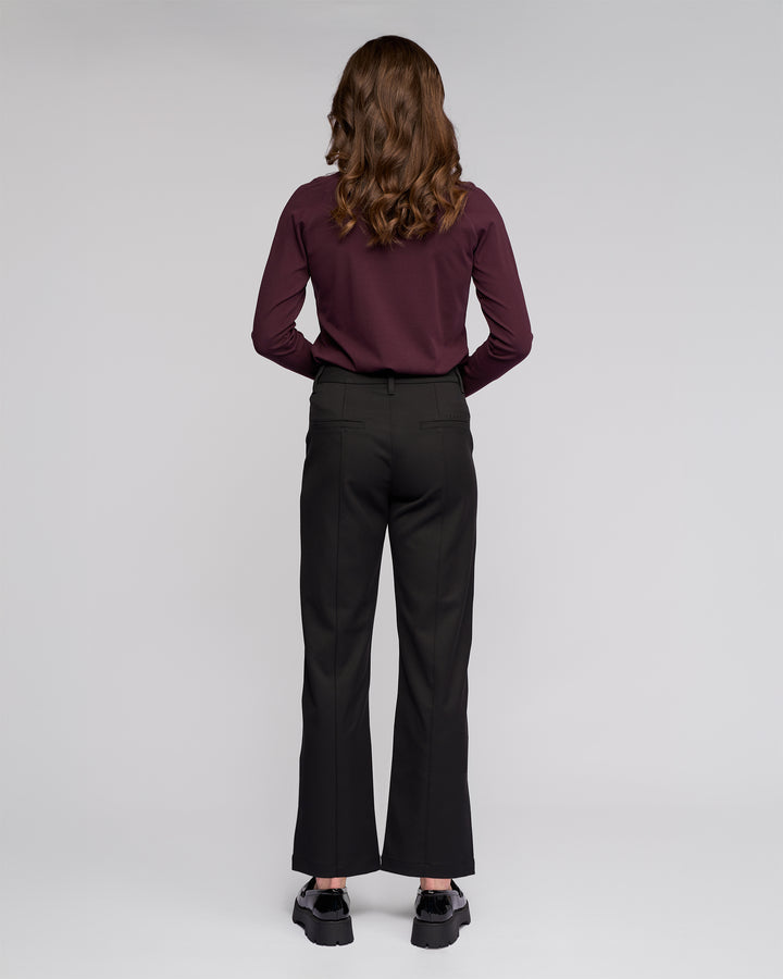 A person with wavy brown hair, dressed in a maroon long-sleeve shirt and Vassalli's Neve Pant in black, paired with black shoes, stands facing away from the camera against a plain light background.