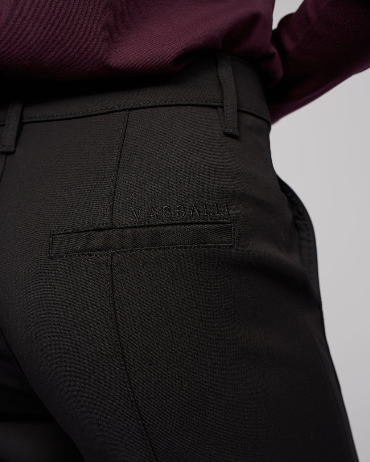Close-up of a person wearing Vassalli’s Neve Pant in black, featuring tailored straight legs and the brand stitched above the back pocket, styled with a dark burgundy top for a classic look.