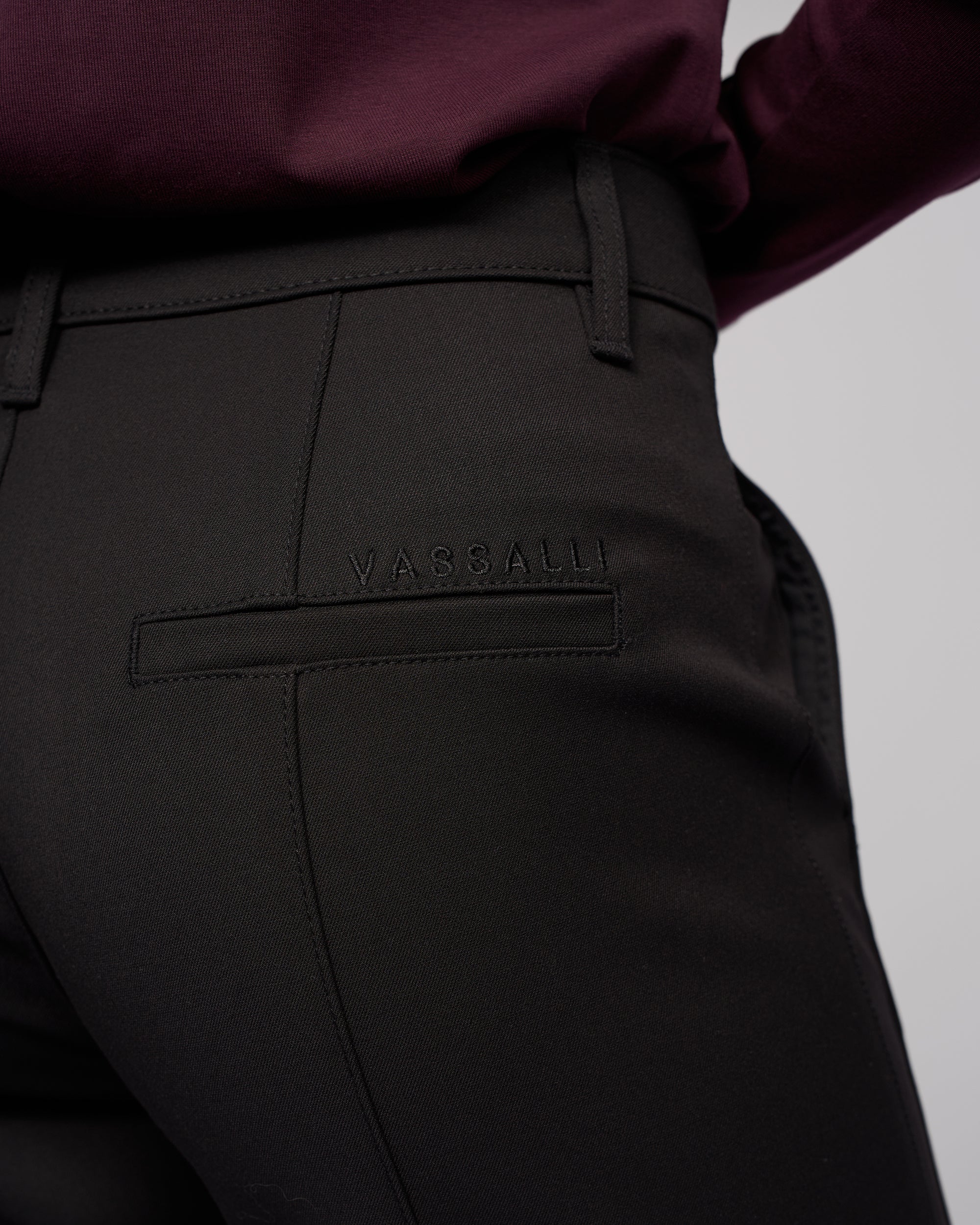 Close-up of a person wearing Vassalli’s Neve Pant in black, featuring tailored straight legs and the brand stitched above the back pocket, styled with a dark burgundy top for a classic look.