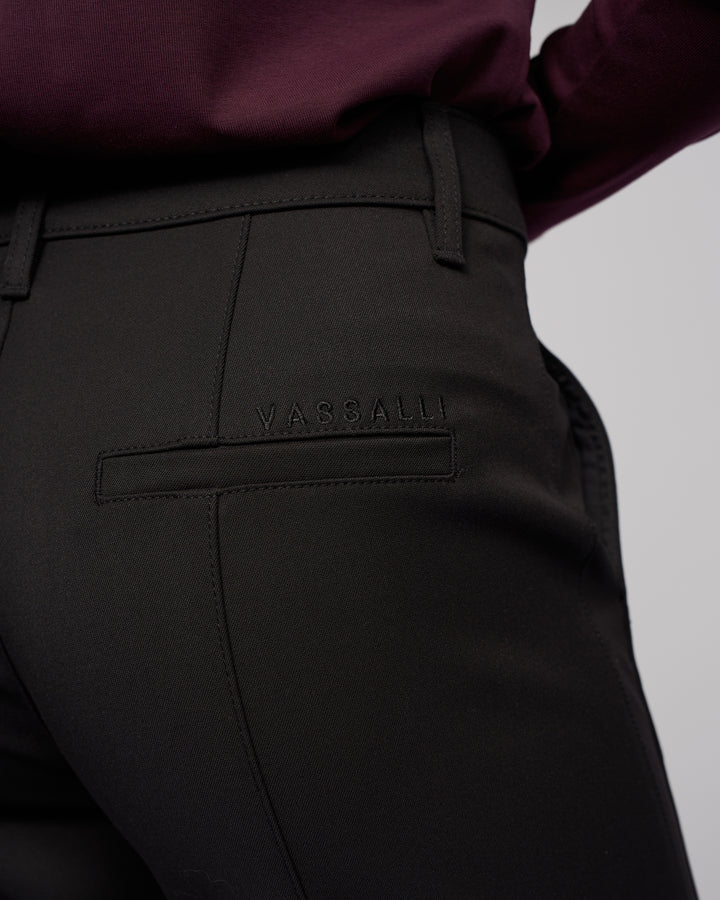 Close-up of a person wearing Vassalli’s Neve Pant in black, featuring tailored straight legs and the brand stitched above the back pocket, styled with a dark burgundy top for a classic look.
