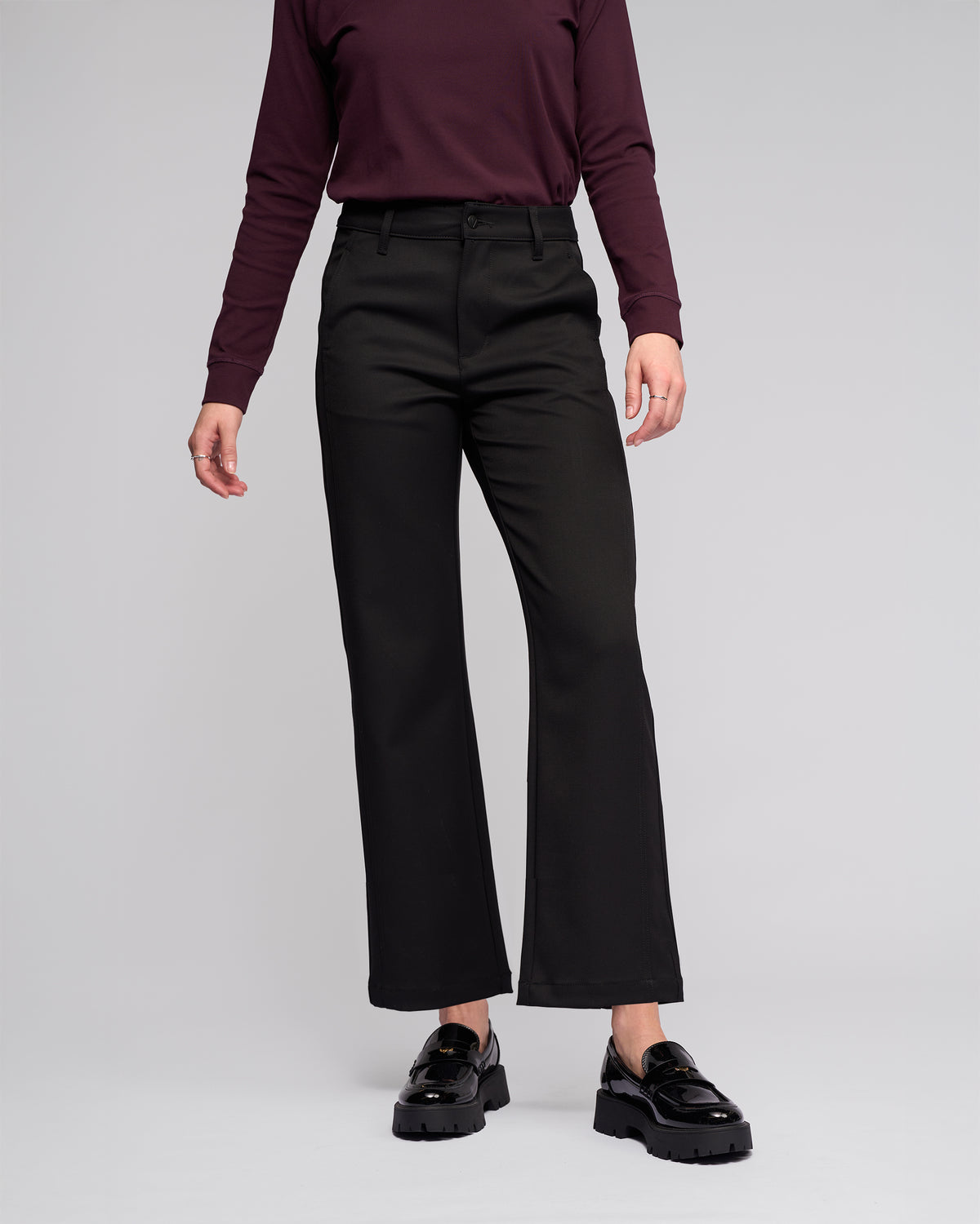 A person models the Vassalli Neve Pant in black with a long-sleeve maroon top and shiny black loafers against a plain light gray background. Their head is not shown in the image.