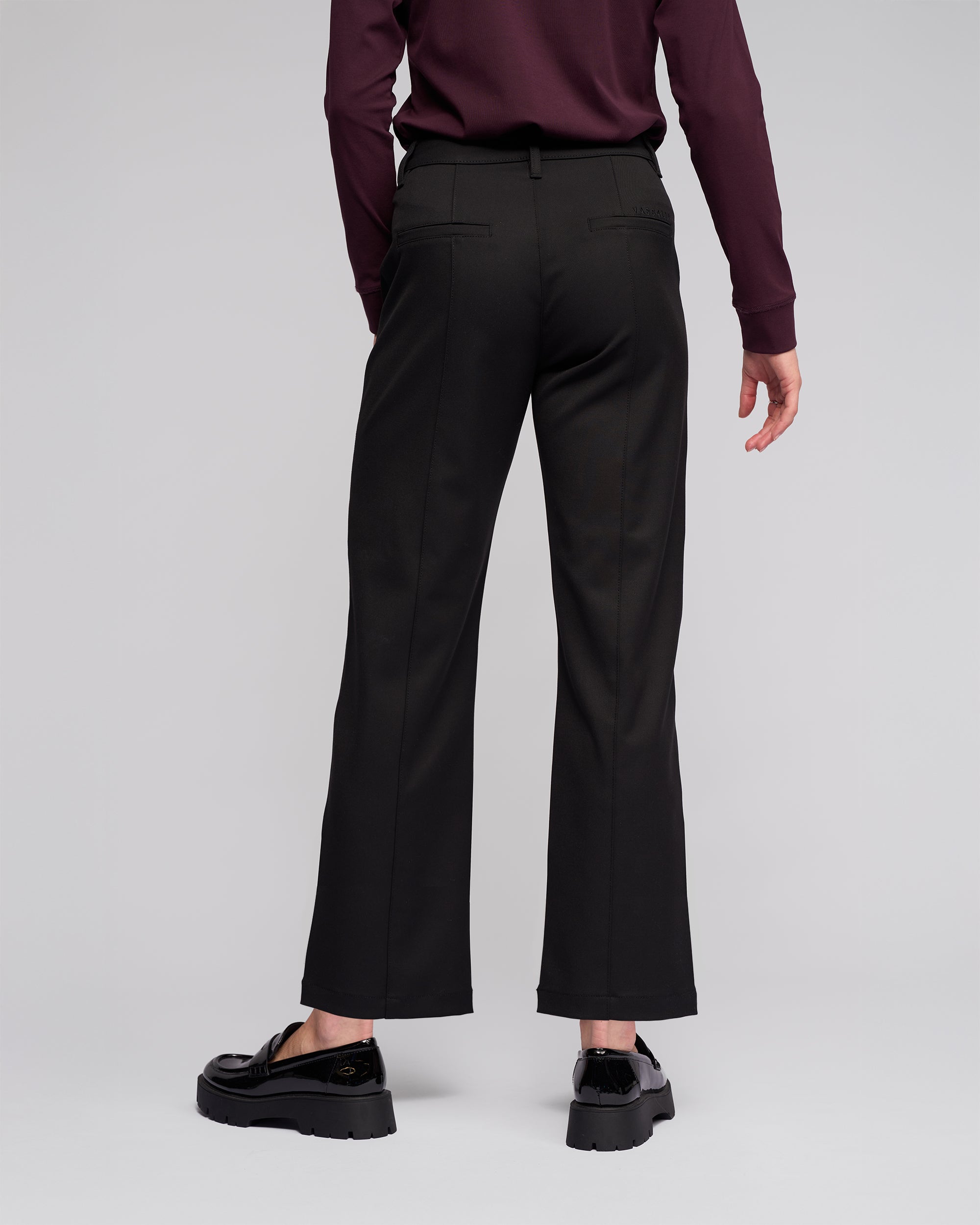 A person stands facing away against a light gray background, wearing Vassalli's Neve Pant in black, a long-sleeved maroon top, and black loafers.