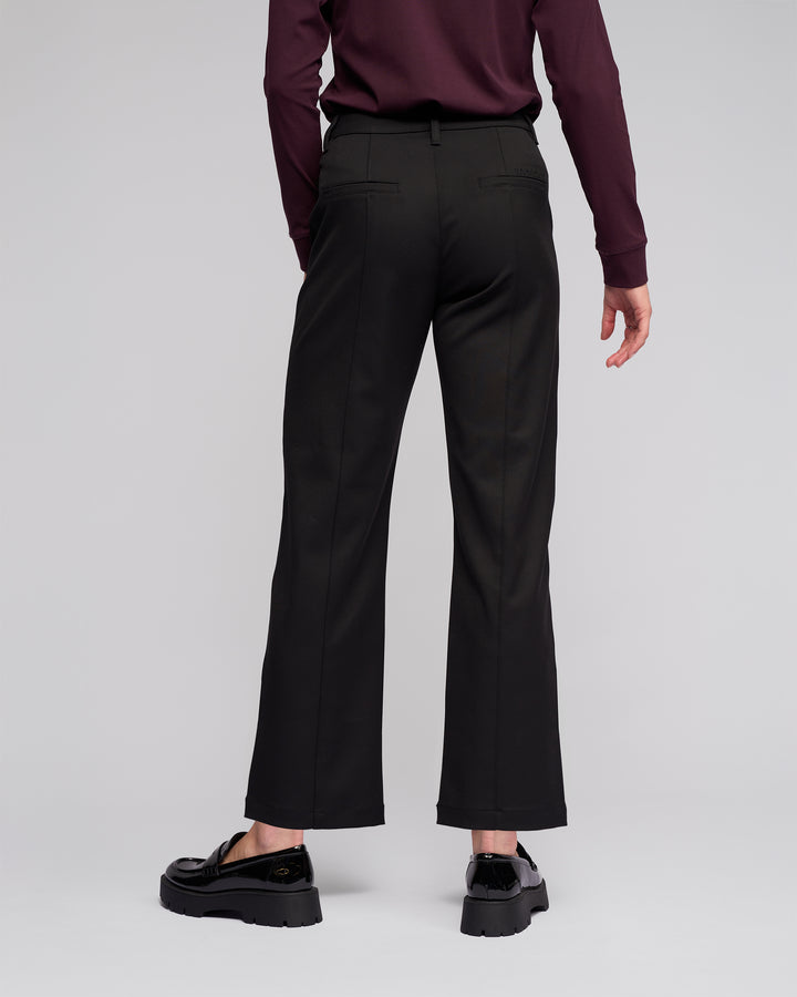 A person stands facing away against a light gray background, wearing Vassalli's Neve Pant in black, a long-sleeved maroon top, and black loafers.