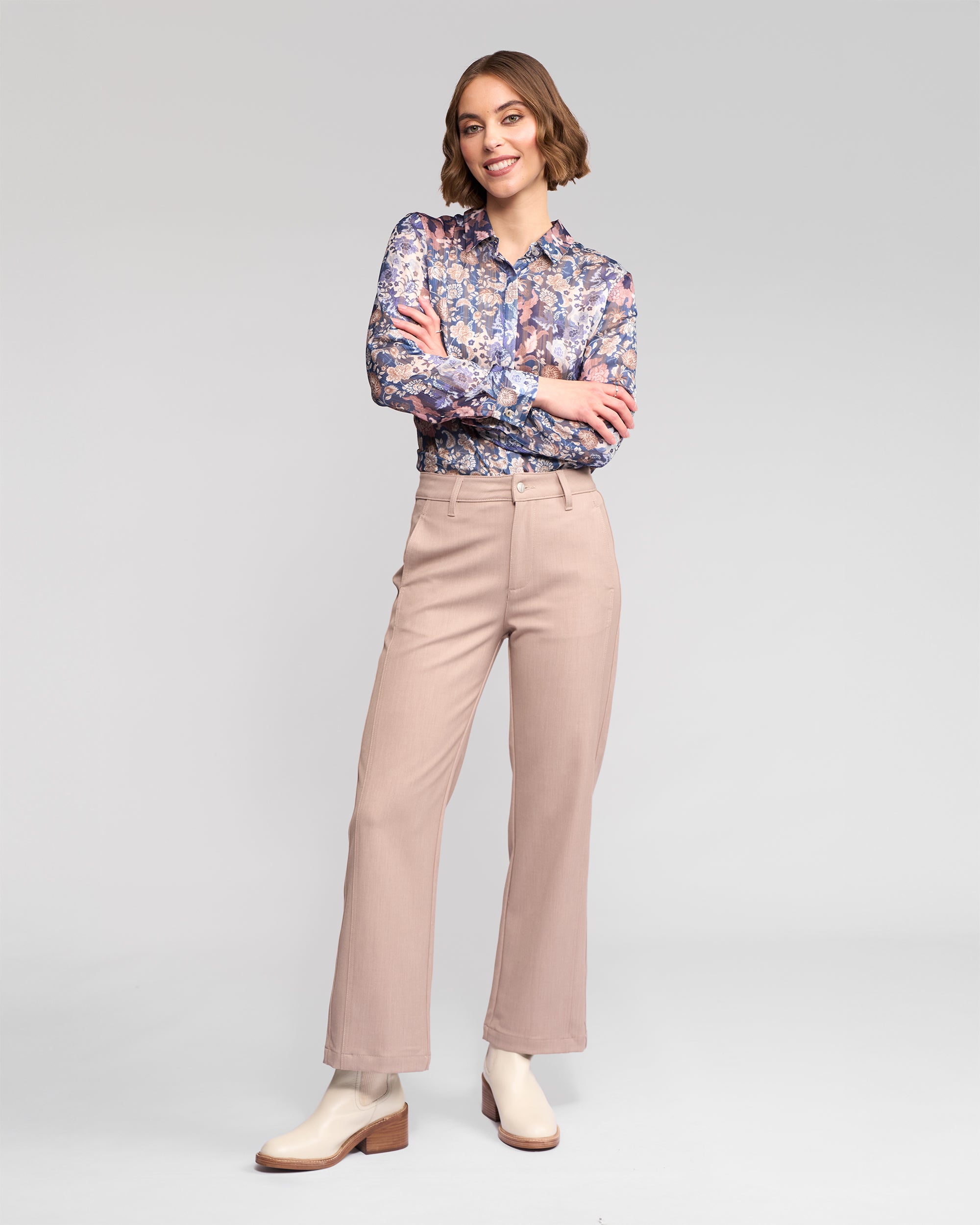A woman smiles with arms crossed, wearing a floral long-sleeve blouse, Vassalli Neve Pant in Oyster, and cream ankle boots against a plain light gray background.